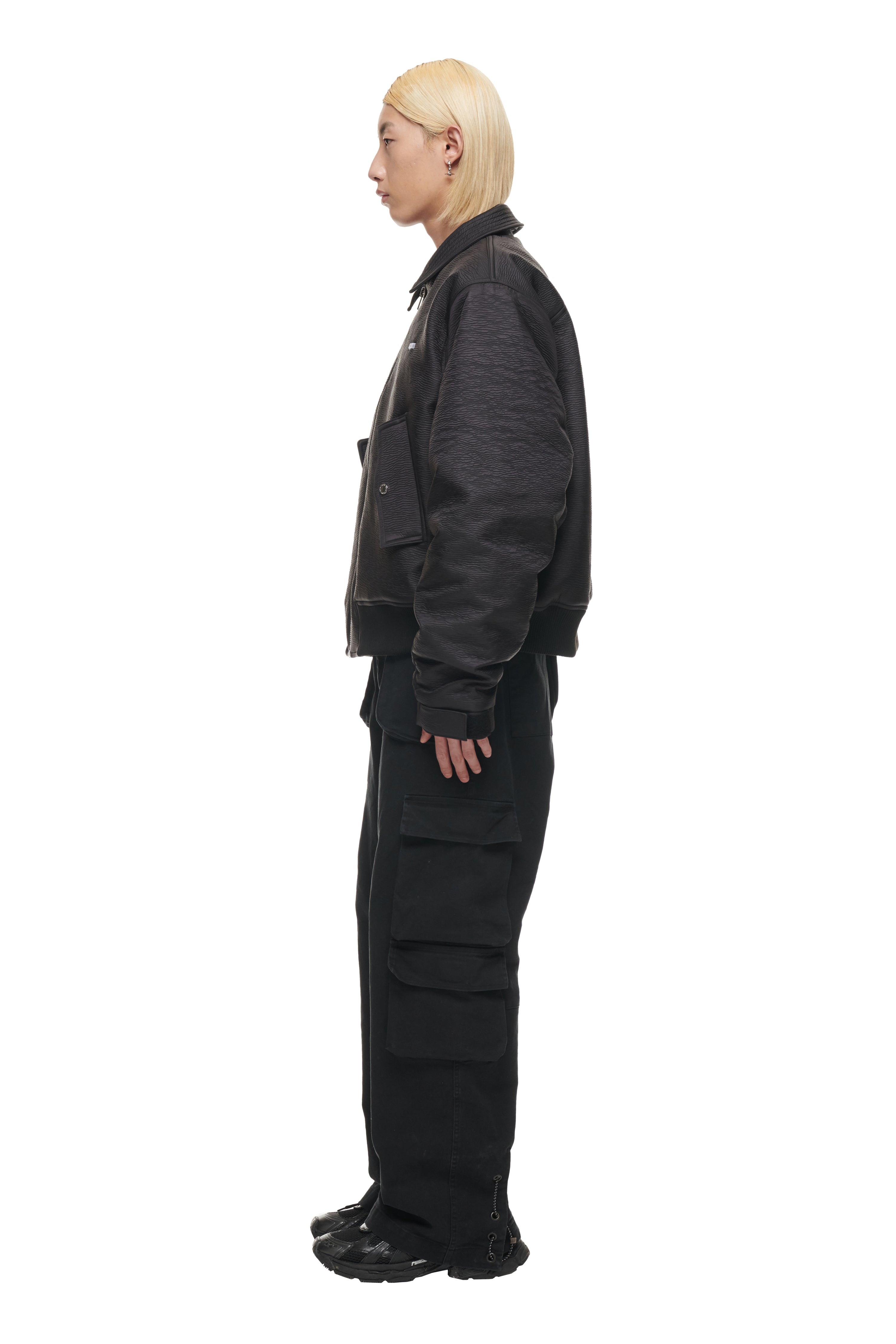 Person with straight, platinum blonde hair in profile wears the 6PM BOMBER JACKET BLACK, black cargo pants with large side pockets, and black sneakers against a plain white background.