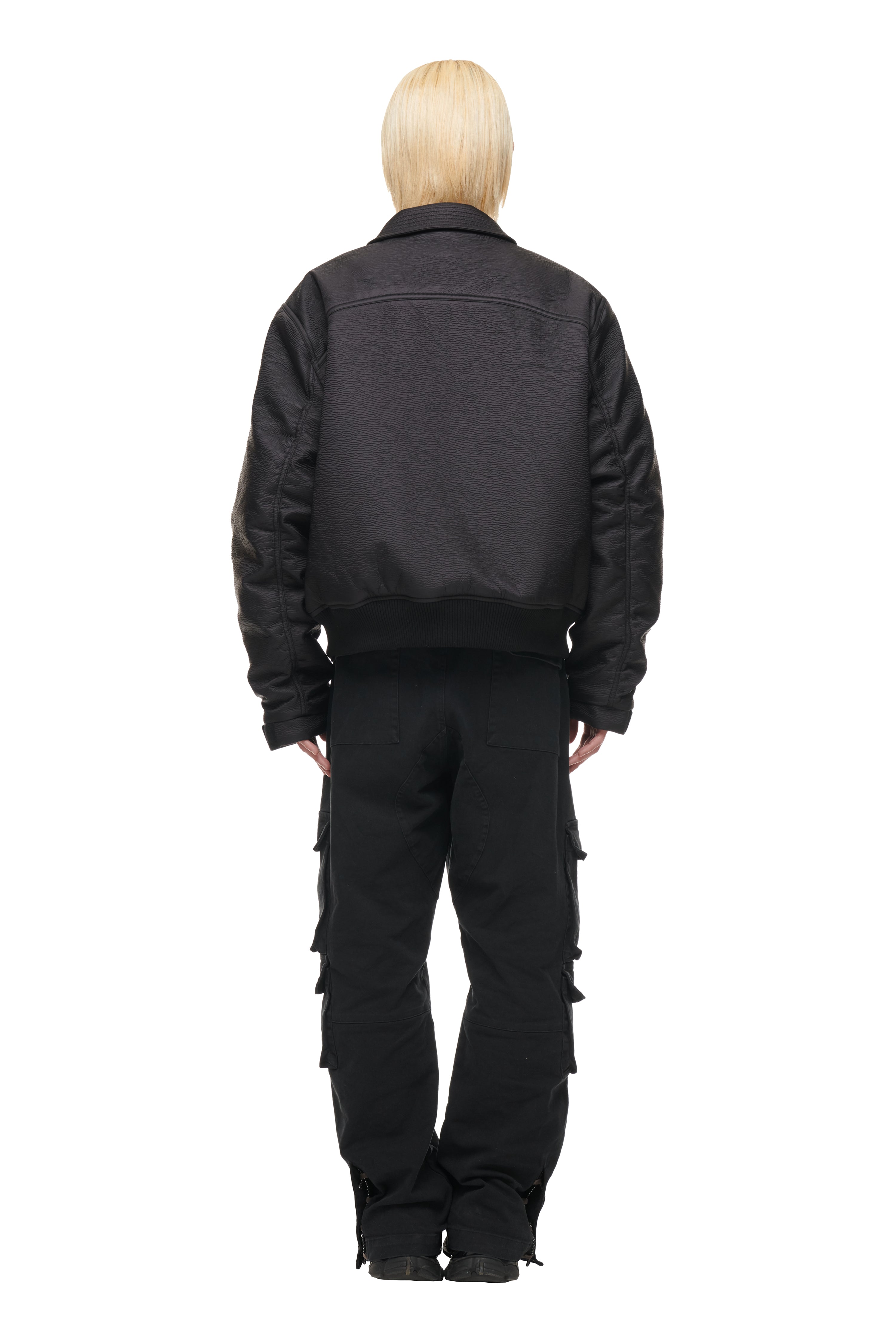 A person with short, light blonde hair models the 6PM BOMBER JACKET BLACK, paired with black cargo pants, standing against a plain white background.