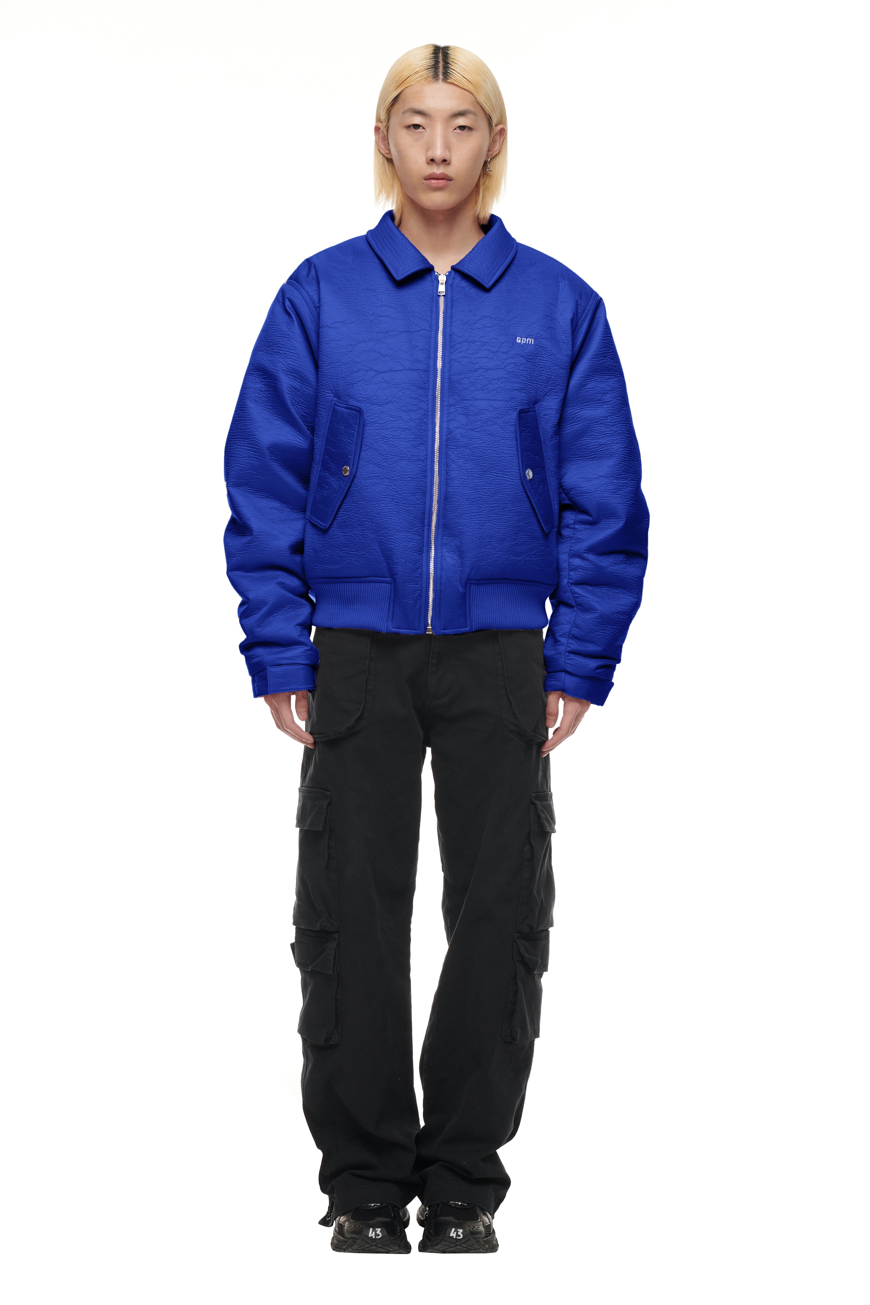 A person with straight platinum blond hair wears the 6PM BOMBER JACKET BLUE, paired with black cargo pants with pockets, standing facing forward against a plain white background.