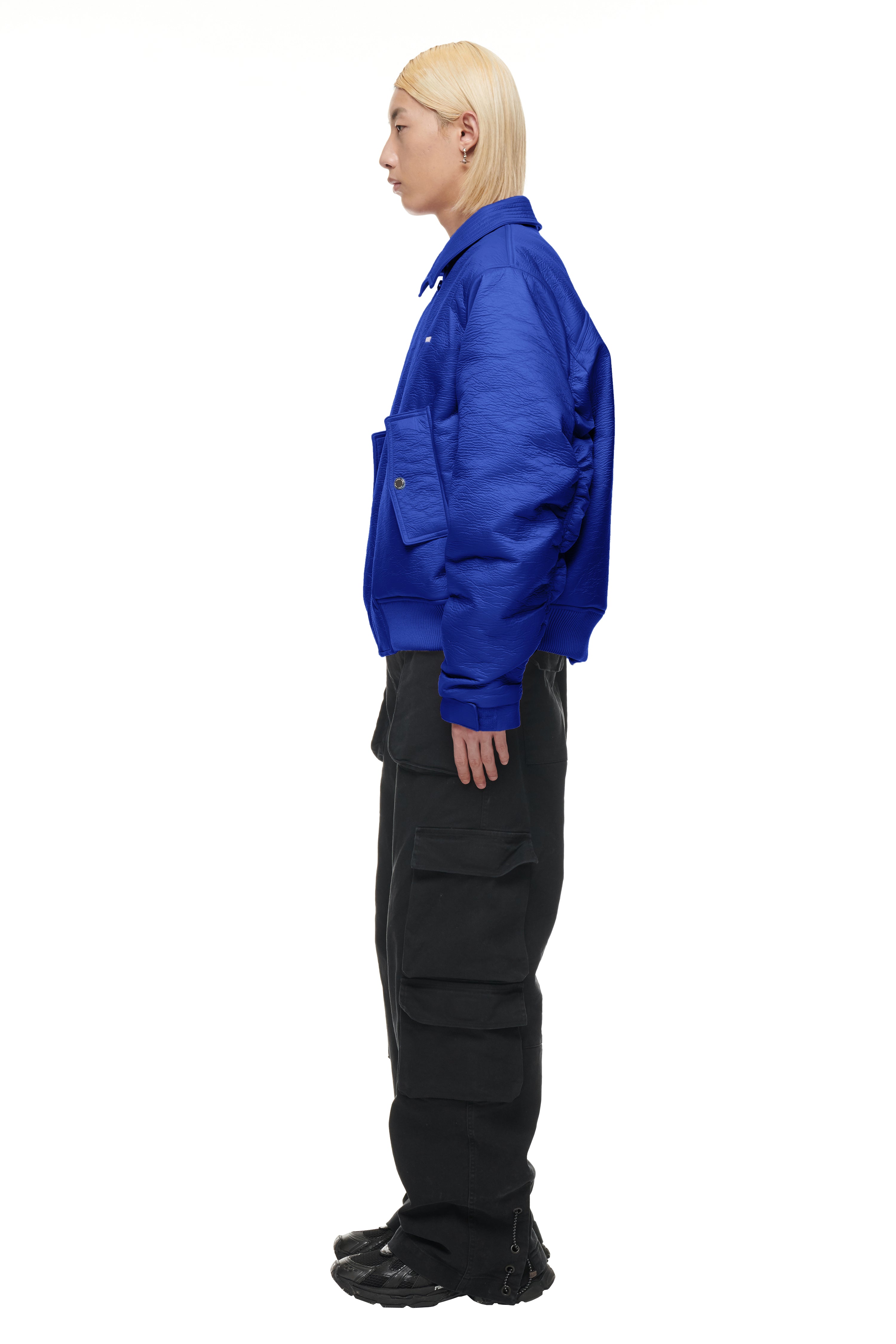 A person with straight platinum blond hair stands in profile against a plain white background, wearing the 6PM BOMBER JACKET BLUE, black cargo pants with large pockets, and black sneakers.