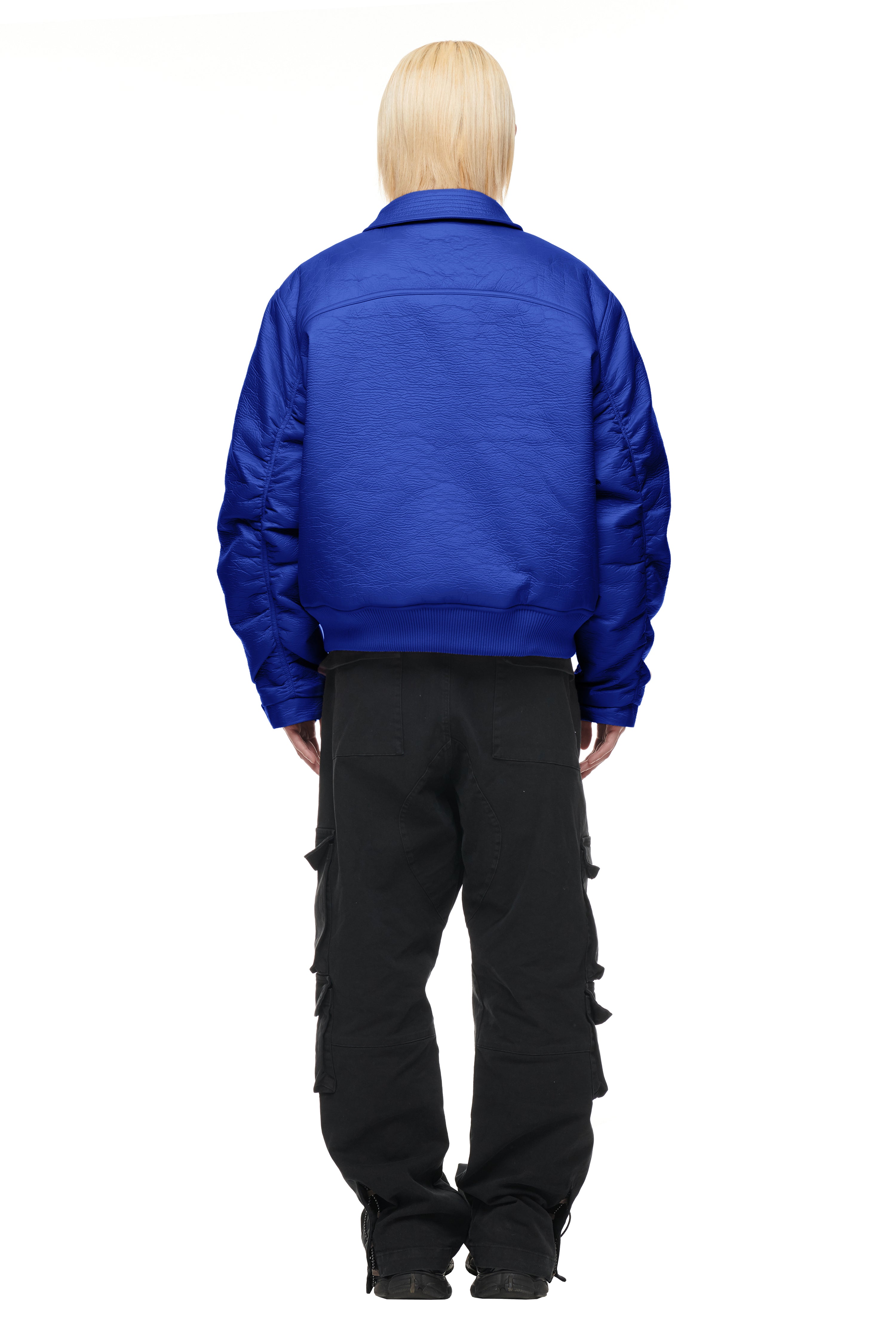 A person with straight, light blonde hair faces away, wearing the 6PM BOMBER JACKET BLUE and black cargo pants with multiple pockets, set against a plain white background.