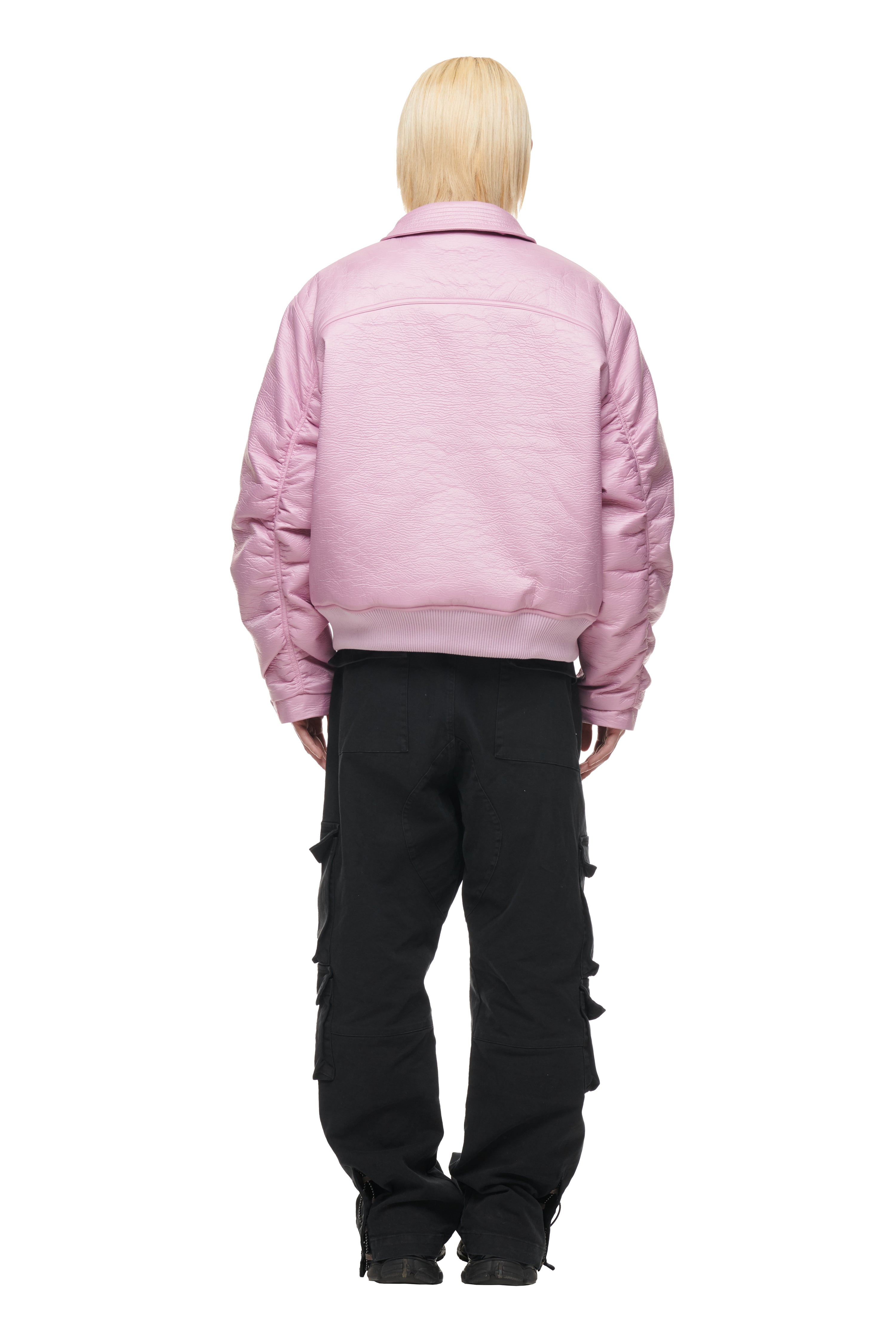 A person with straight, light blonde hair stands facing away, wearing the 6PM BOMBER JACKET PINK and loose black cargo pants with multiple pockets, set against a plain white background.