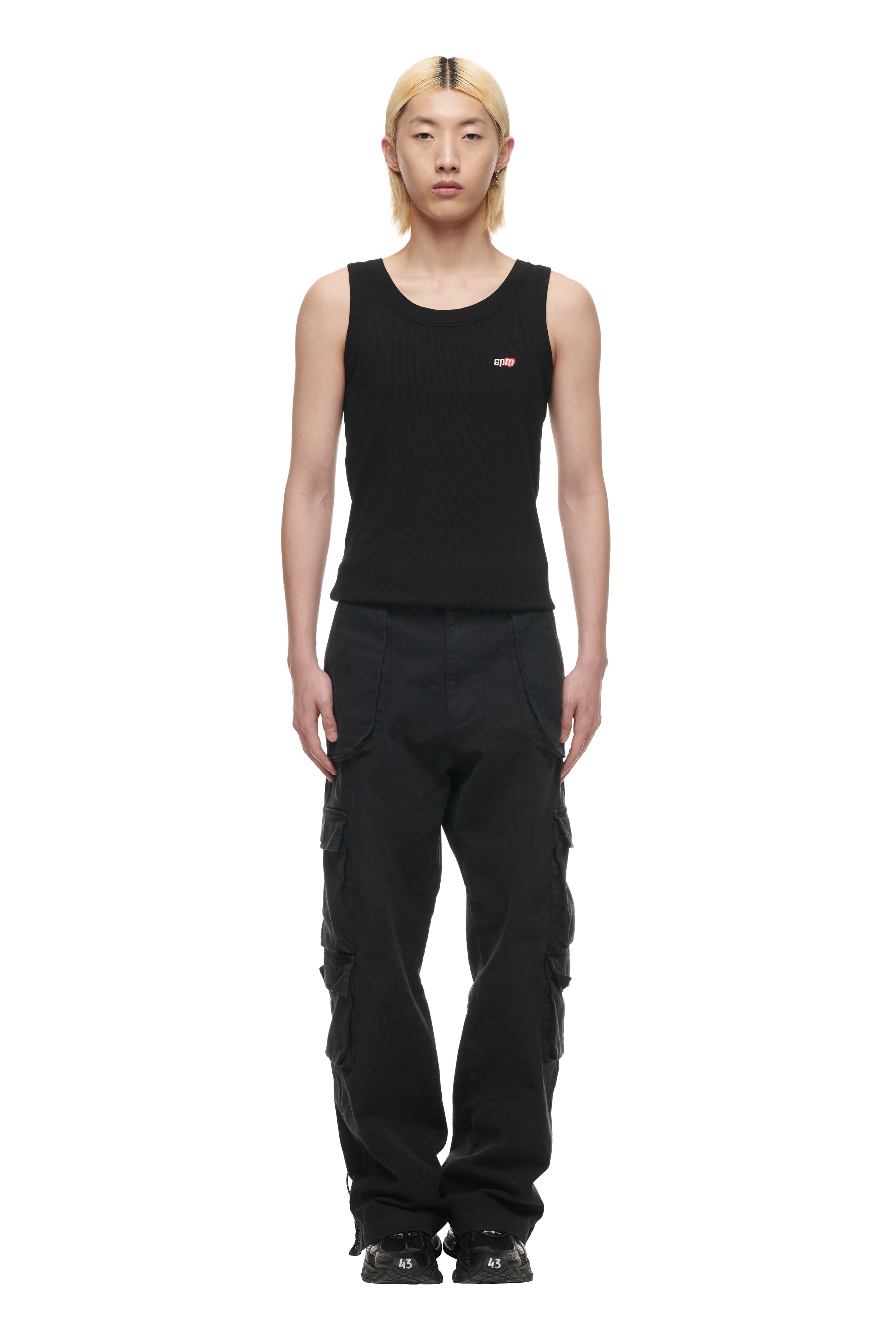 A person with straight blond hair wears the 6PM TANK TOP BLACK (2-PACK), black cargo pants with side pockets, and black sneakers, standing against a plain white background.