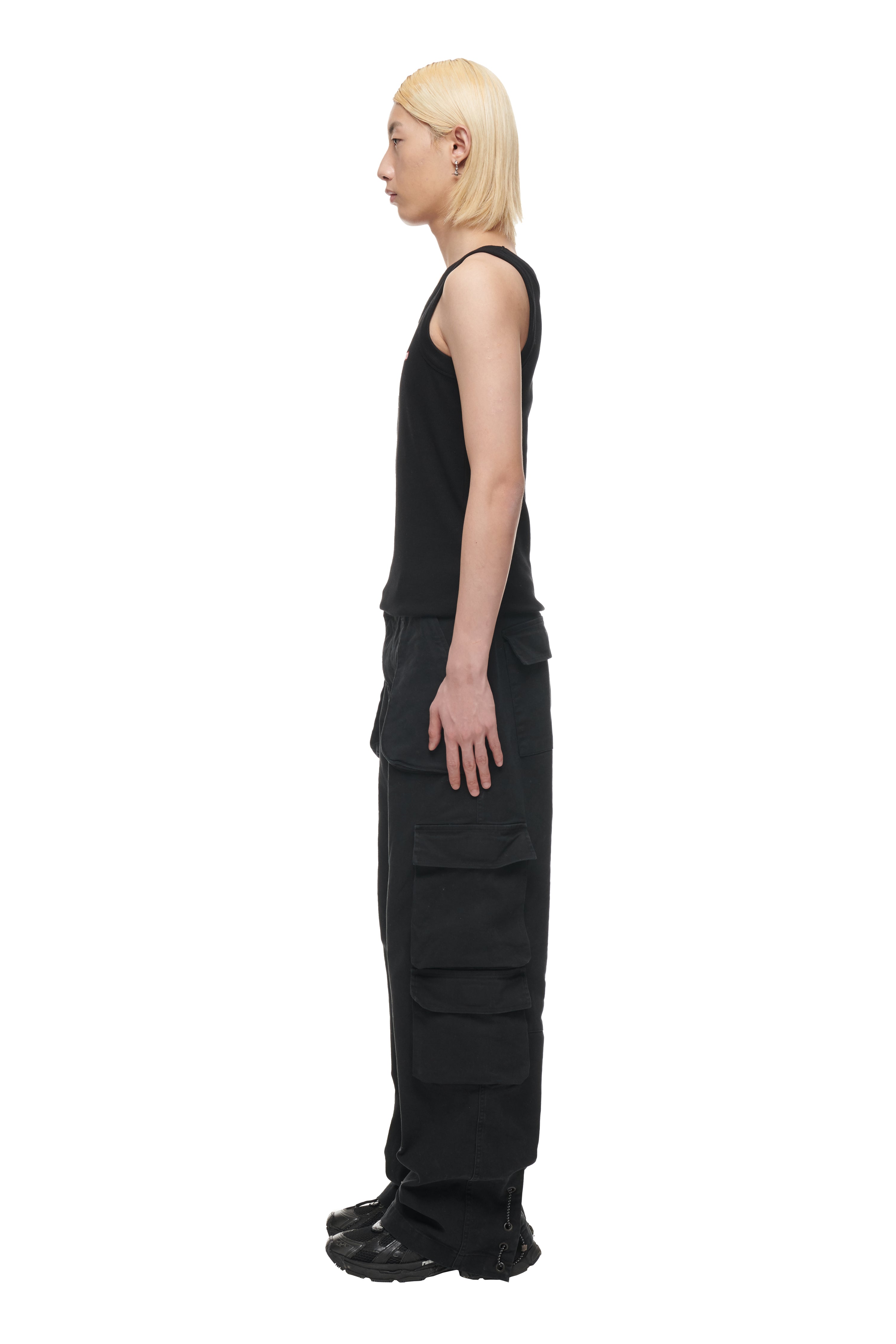 A person with straight, shoulder-length blond hair stands in profile wearing the 6PM TANK TOP BLACK (2-PACK), black cargo pants with large pockets, and black shoes, set against a white background.