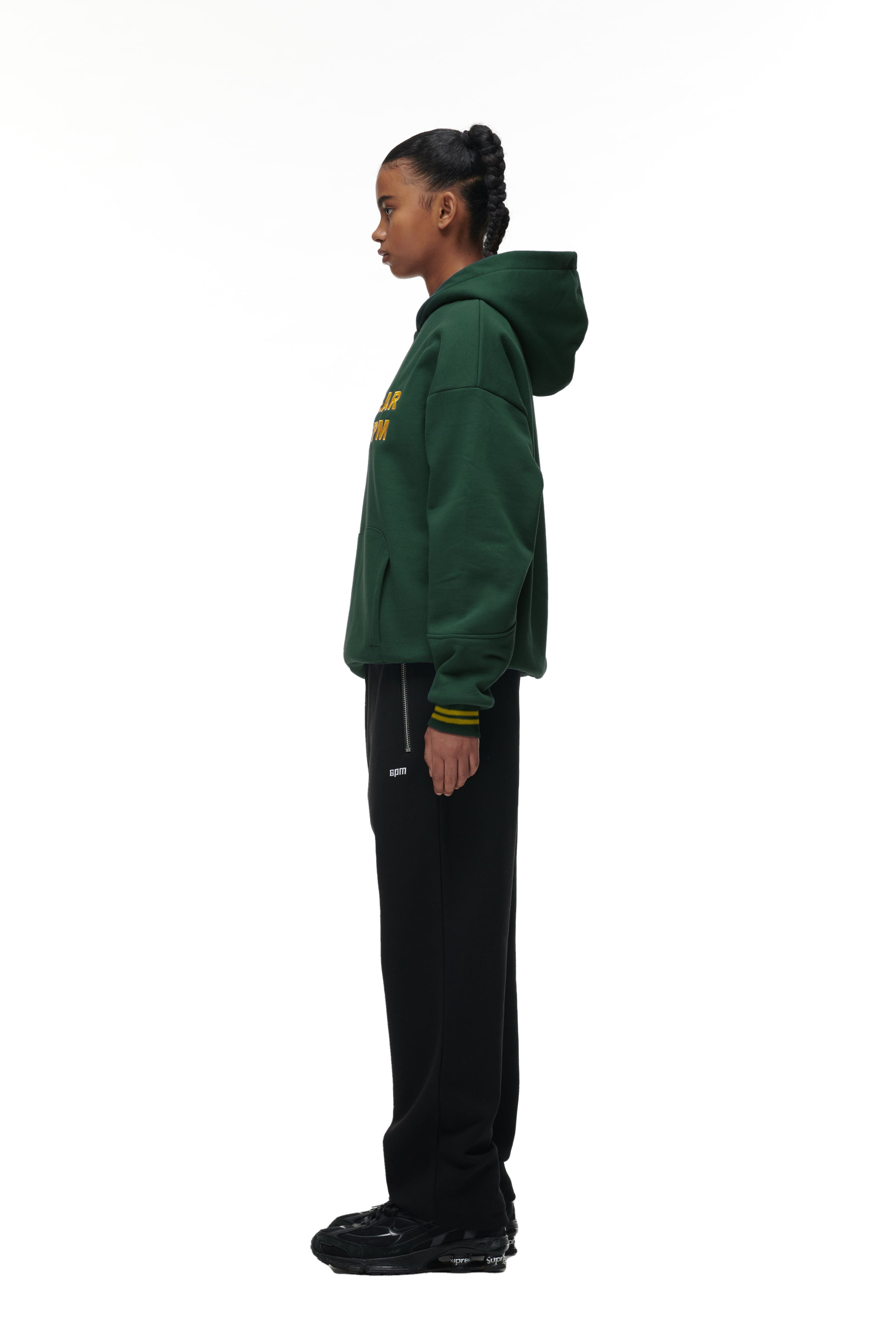 A person stands in profile against a white background, wearing the 6PM ONLY WEAR HOODIE GREEN, paired with black pants and black sneakers. Their hair is styled in a neat braid.