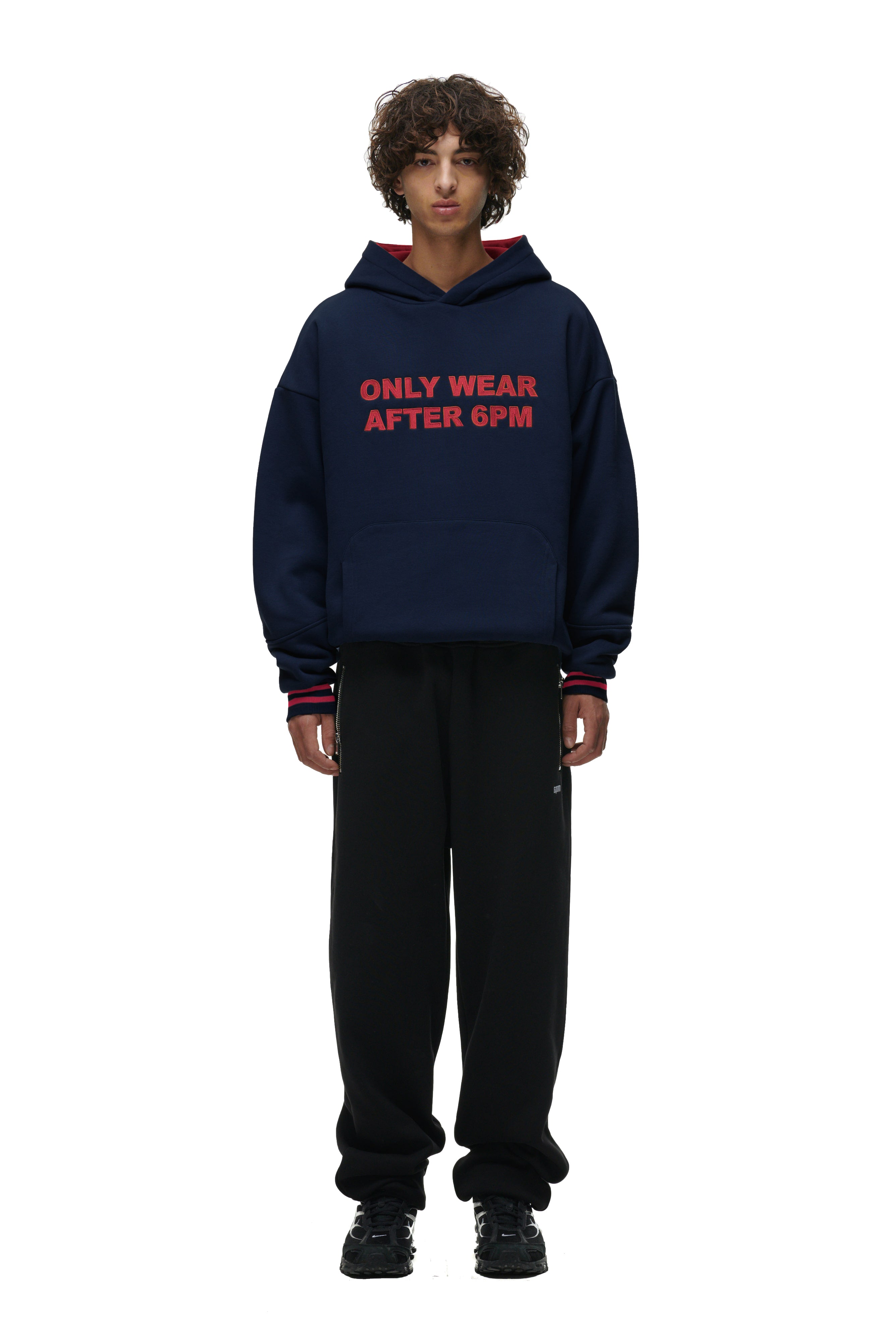A person with curly hair wears the 6PM ONLY WEAR HOODIE NAVY, featuring red text, paired with black pants and black sneakers, standing against a white background.
