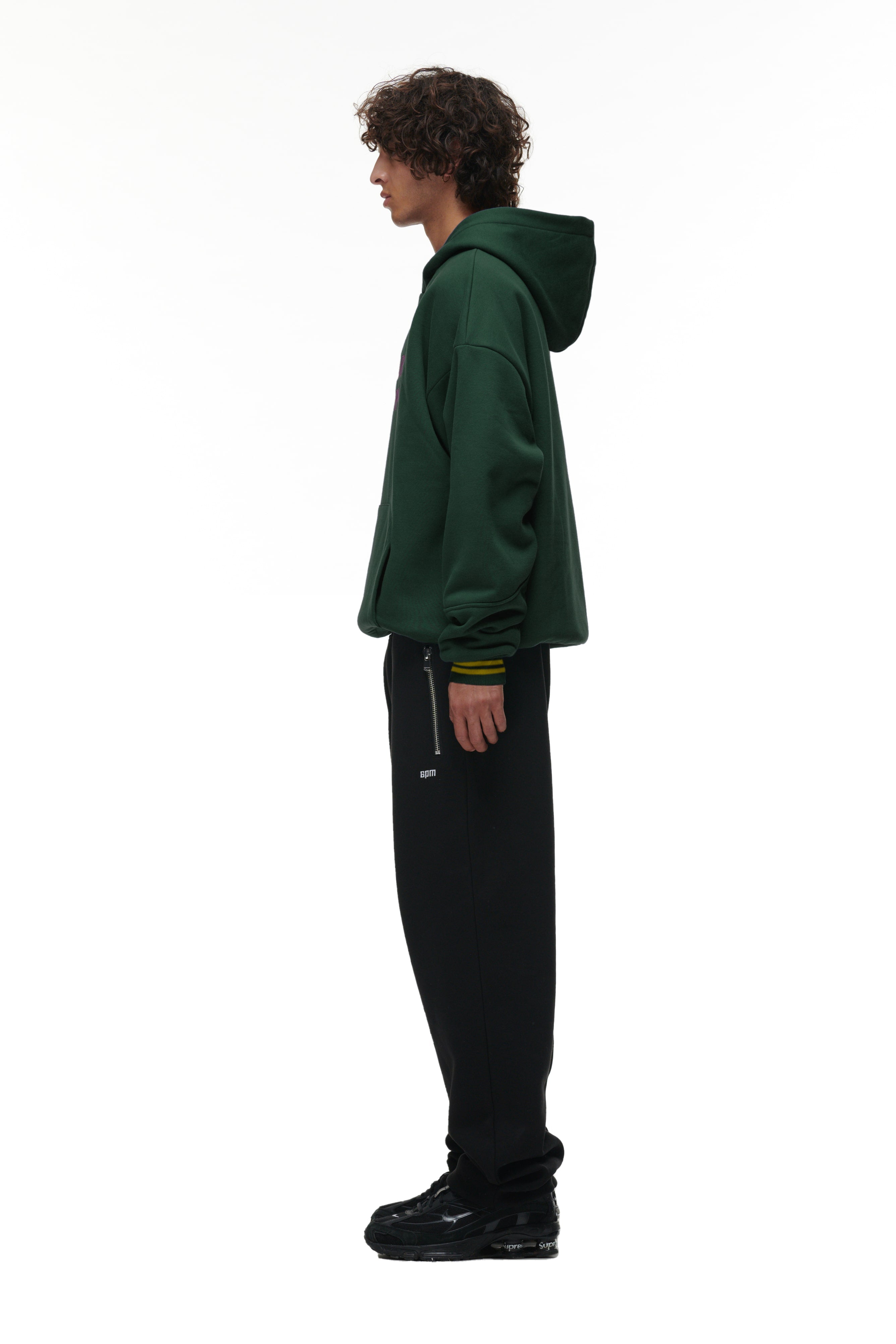 A person with curly hair stands in profile against a white background, wearing the 6PM ONLY WEAR HOODIE GREEN, black pants with a zipper pocket, and black shoes.