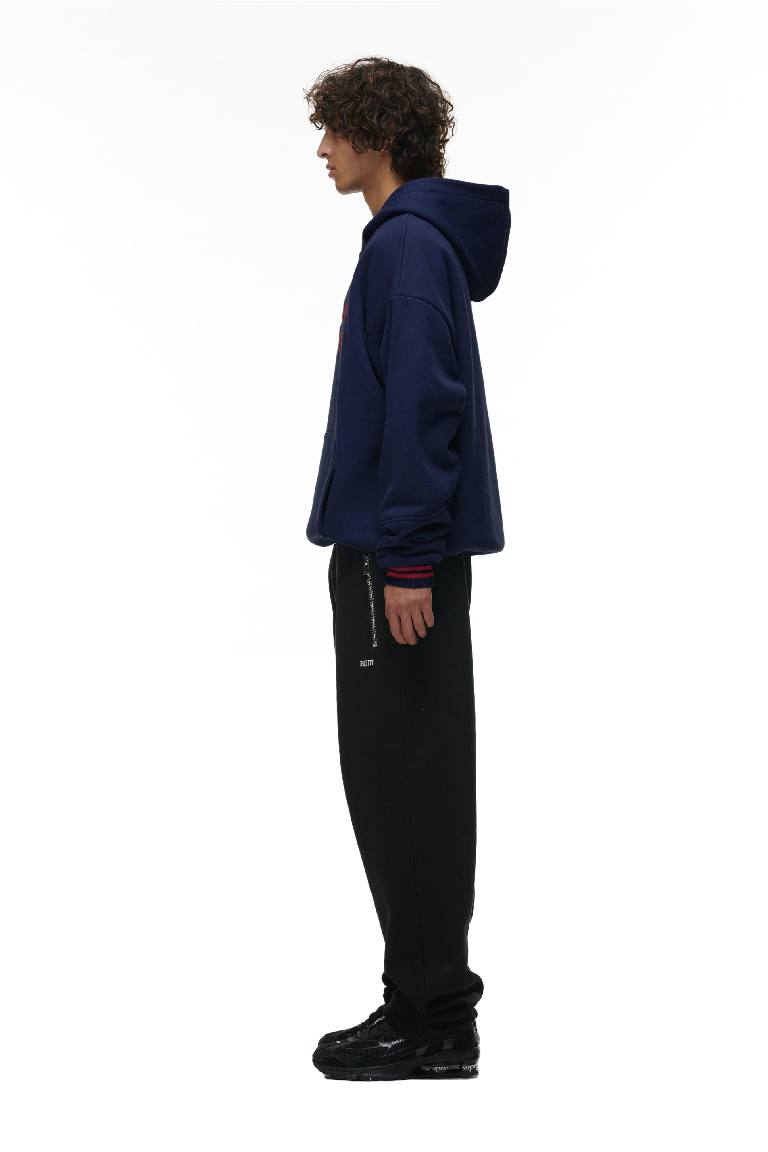 A person with curly hair stands in profile against a white background, wearing the 6PM ONLY WEAR HOODIE NAVY, black pants with a visible zipper, and black shoes.