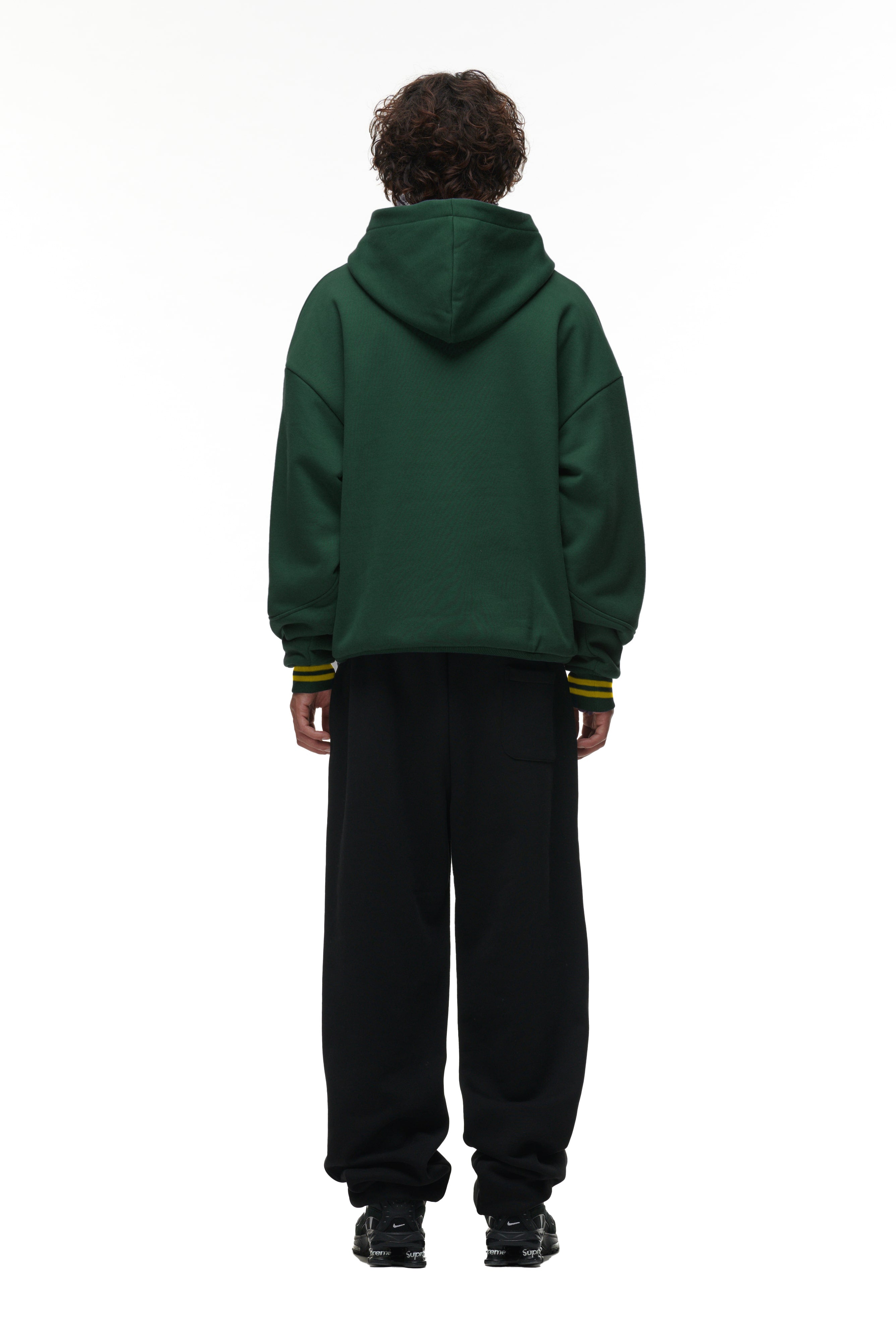 A person stands with their back to the camera, wearing the 6PM ONLY WEAR HOODIE GREEN, paired with black pants and black shoes, against a plain white background.