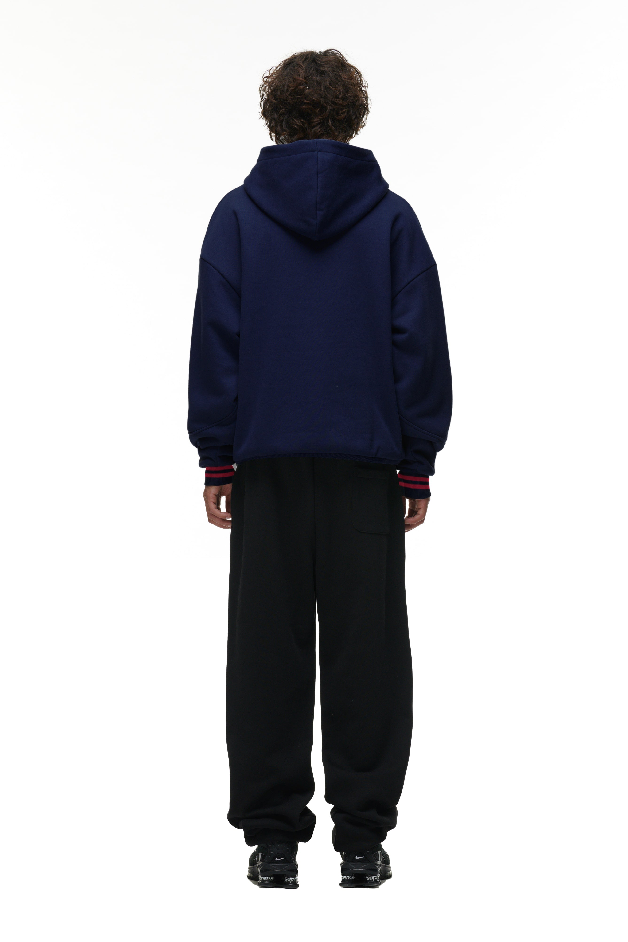 A person stands facing away against a plain white background, wearing the 6PM ONLY WEAR HOODIE NAVY paired with black loose-fitting pants and black shoes.