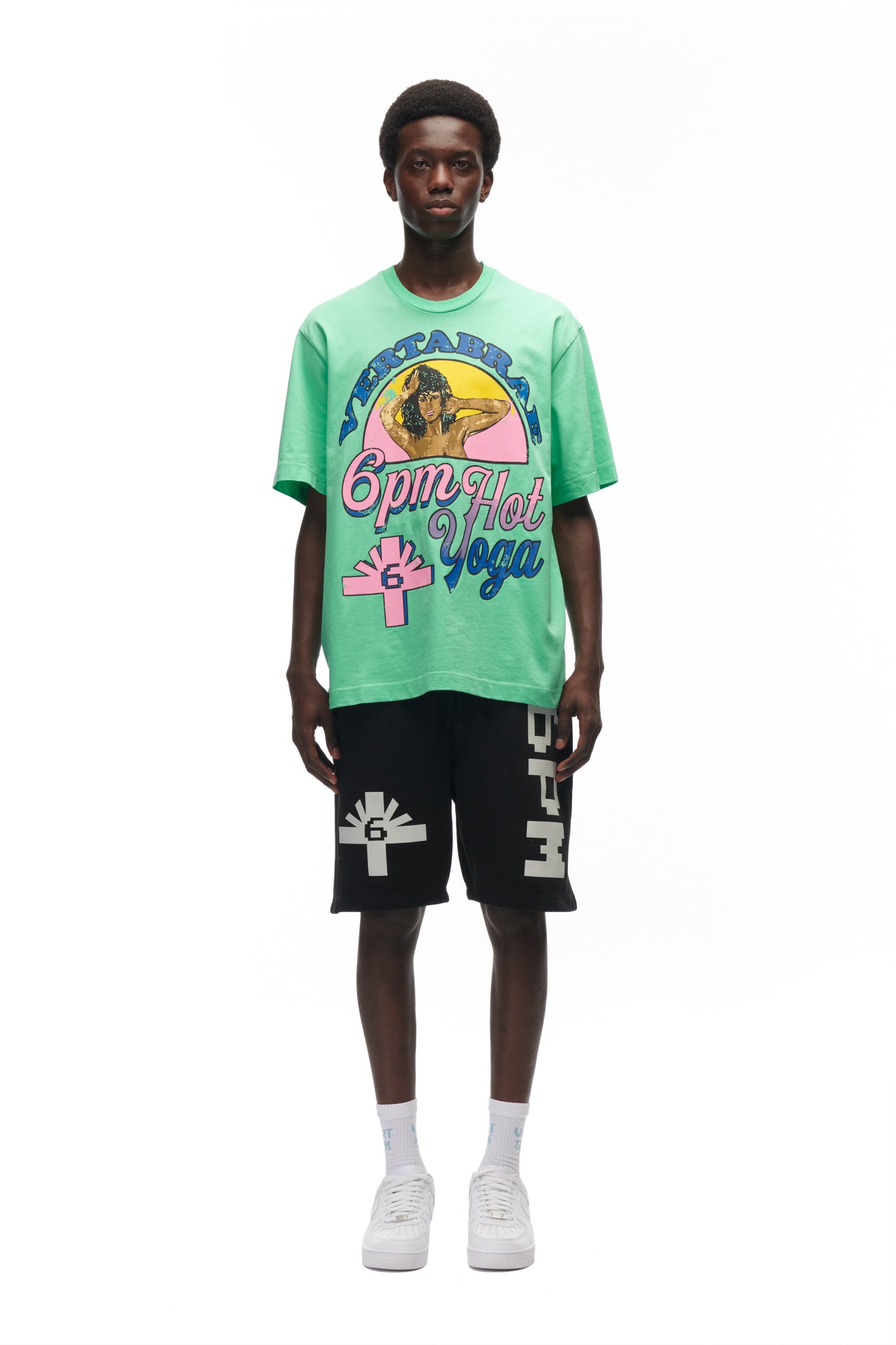 A young man stands facing forward wearing the 6PM HOT YOGA T-SHIRT GREEN, black shorts with white designs, white socks, and white sneakers against a plain white background.