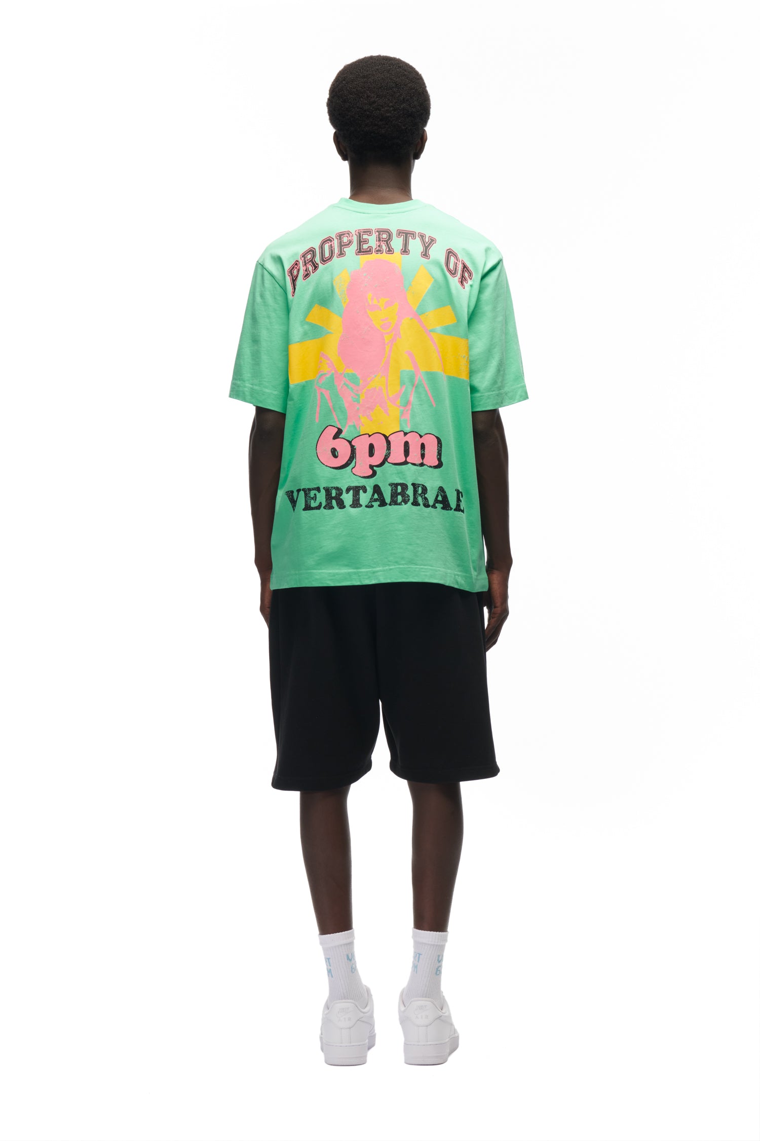A person stands facing away against a white background, wearing the 6PM HOT YOGA T-SHIRT GREEN featuring colorful graphics and "PROPERTY OF 6pm VERTEBRAE" on the back, paired with black shorts, white socks, and white sneakers.