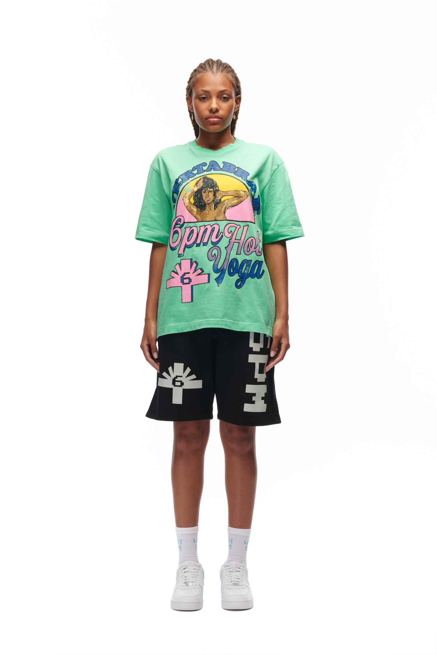A person with braided hair faces forward, wearing the 6PM HOT YOGA T-SHIRT GREEN, black shorts with white designs, white socks, and white sneakers, posed against a plain white background.
