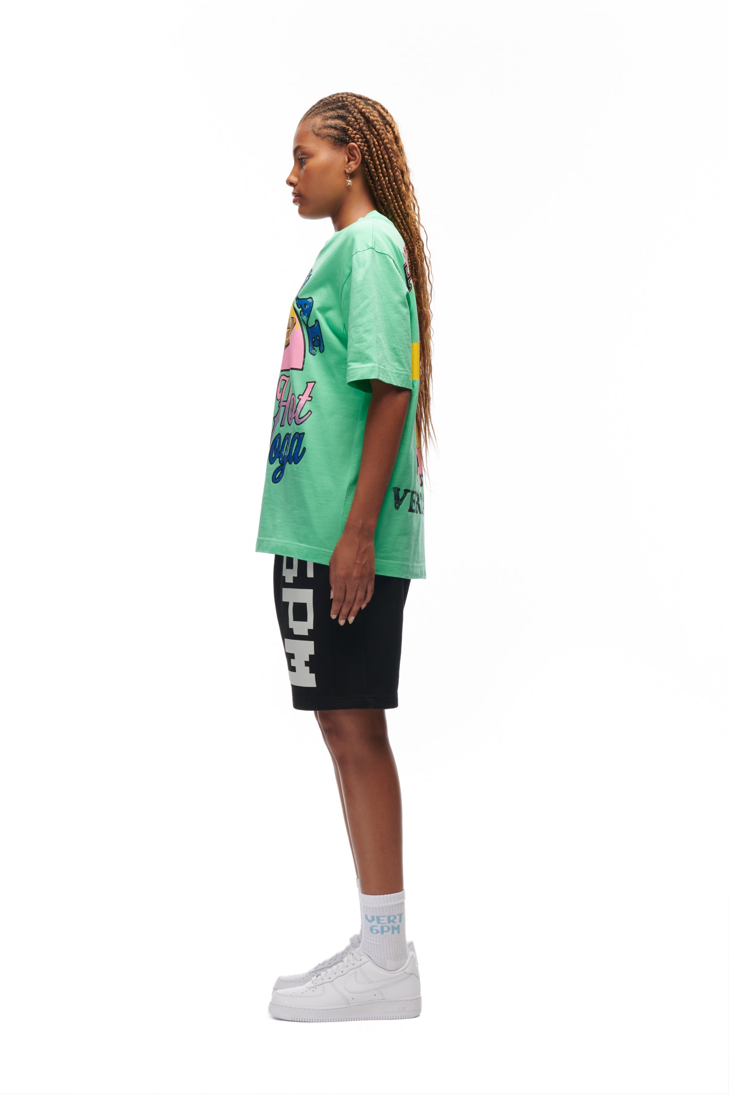 A woman with long braided hair stands in profile against a white background, wearing the 6PM HOT YOGA T-SHIRT GREEN, black shorts with white text, white socks, and white sneakers.