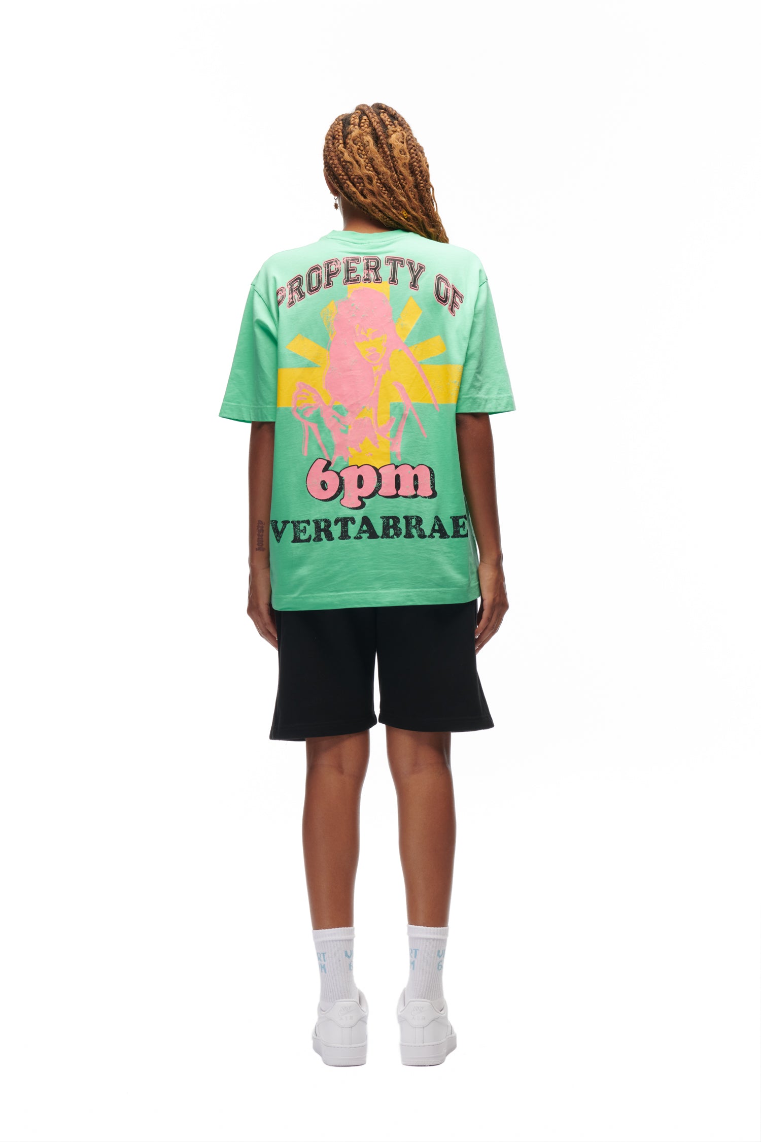 A person with long, braided hair faces away, wearing the 6PM HOT YOGA T-SHIRT GREEN featuring colorful graphics and text, paired with black shorts, white socks, and white sneakers, all set against a white background.