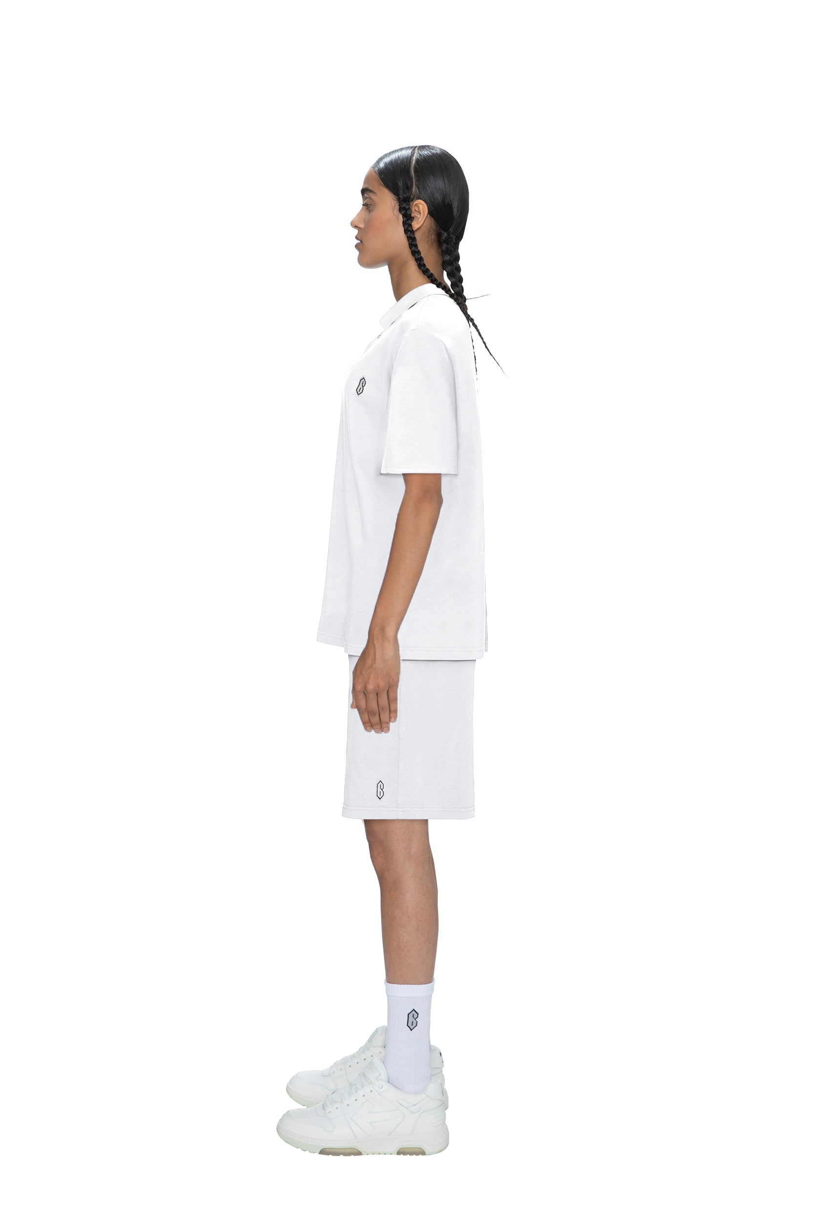 A person with long, braided hair stands in profile against a white background, wearing a 6PM POLO SHIRT WHITE with minimal logos, paired with white shorts, socks, and sneakers.