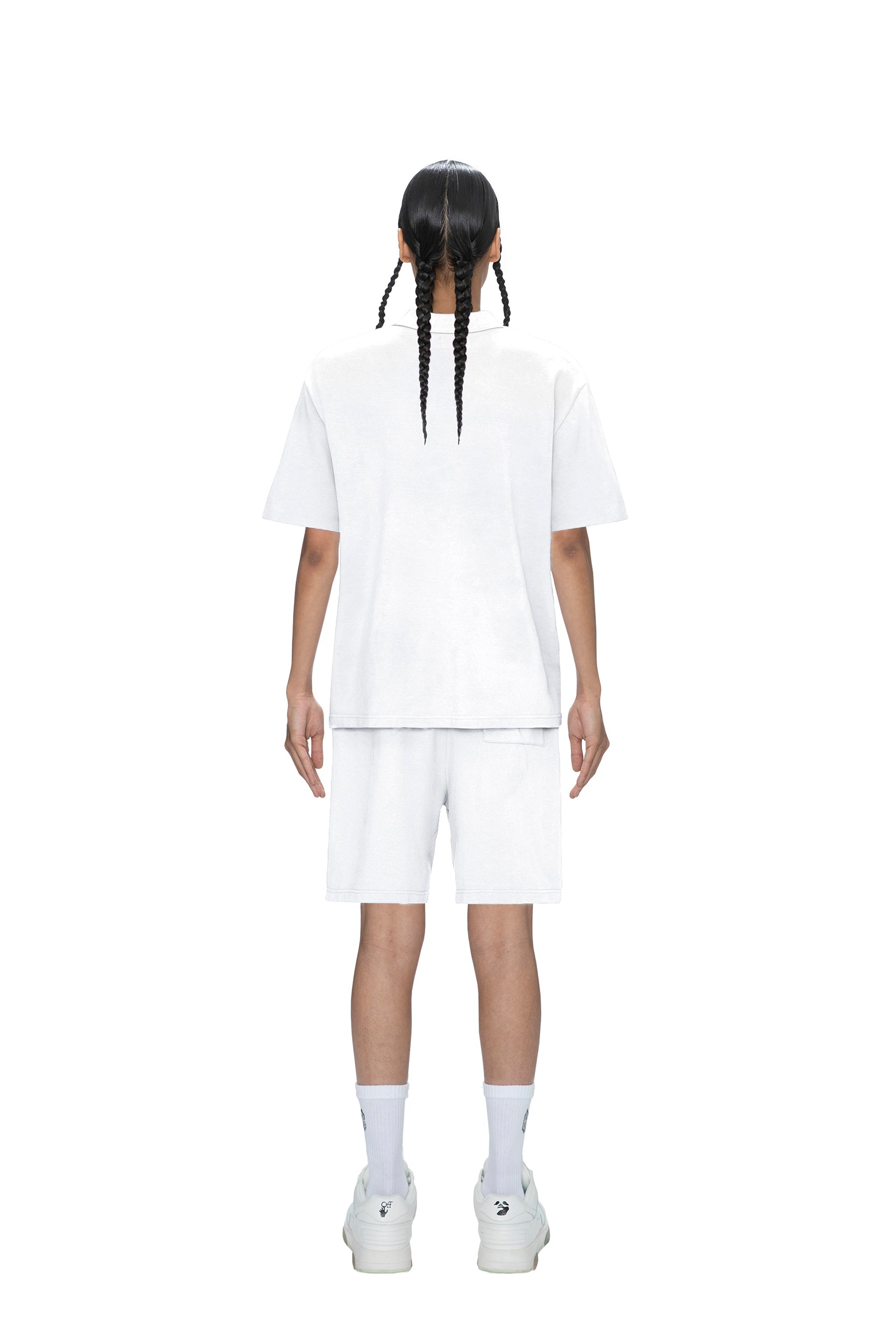 A person with two long braids stands facing away, wearing the 6PM POLO SHIRT WHITE, white shorts, crew socks, and sneakers, all set against a plain white background.