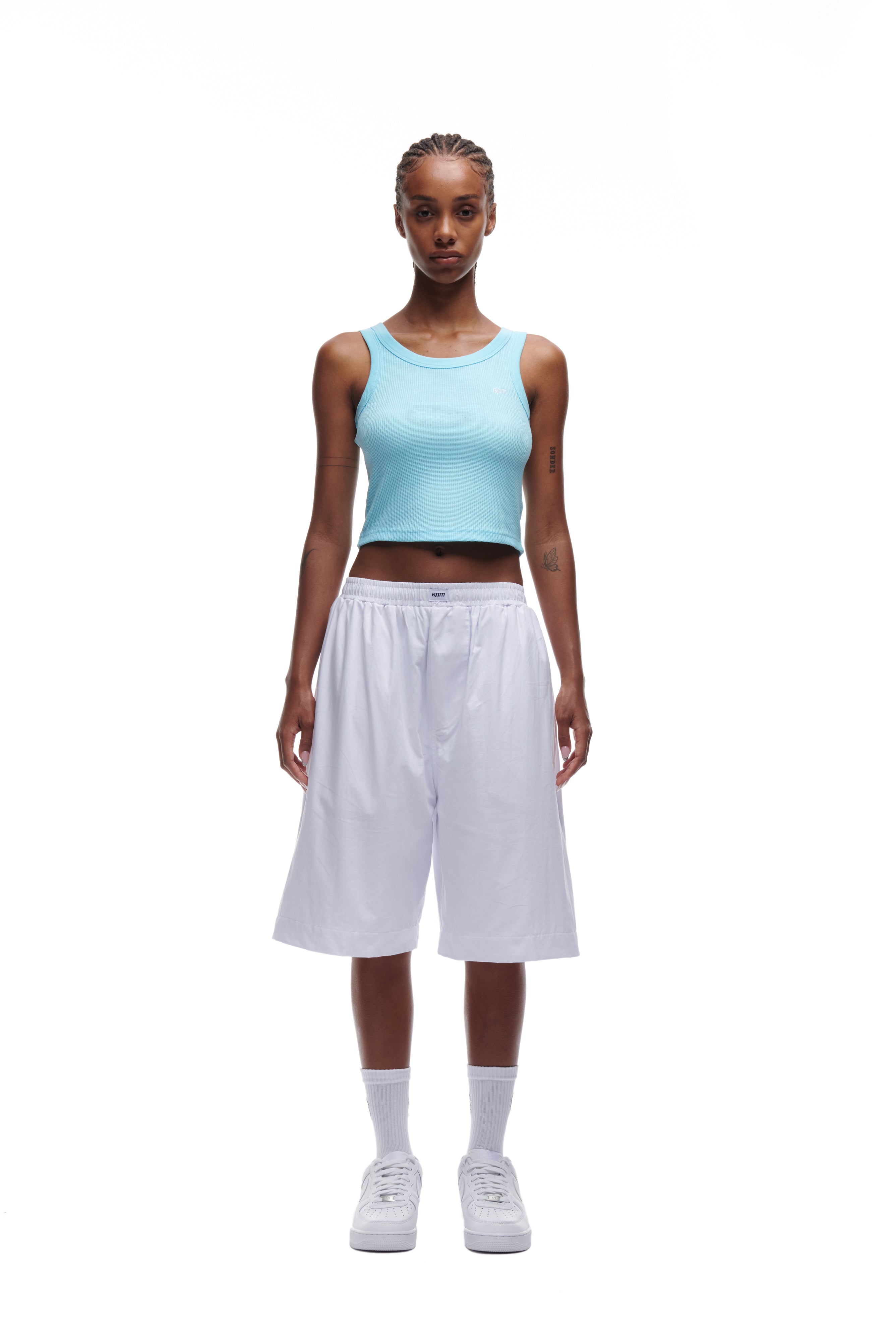 A person stands facing forward, wearing the 6PM WAFFLE KNIT CROP TOP in baby blue, with loose white knee-length shorts, white socks, and white sneakers against a plain white background.