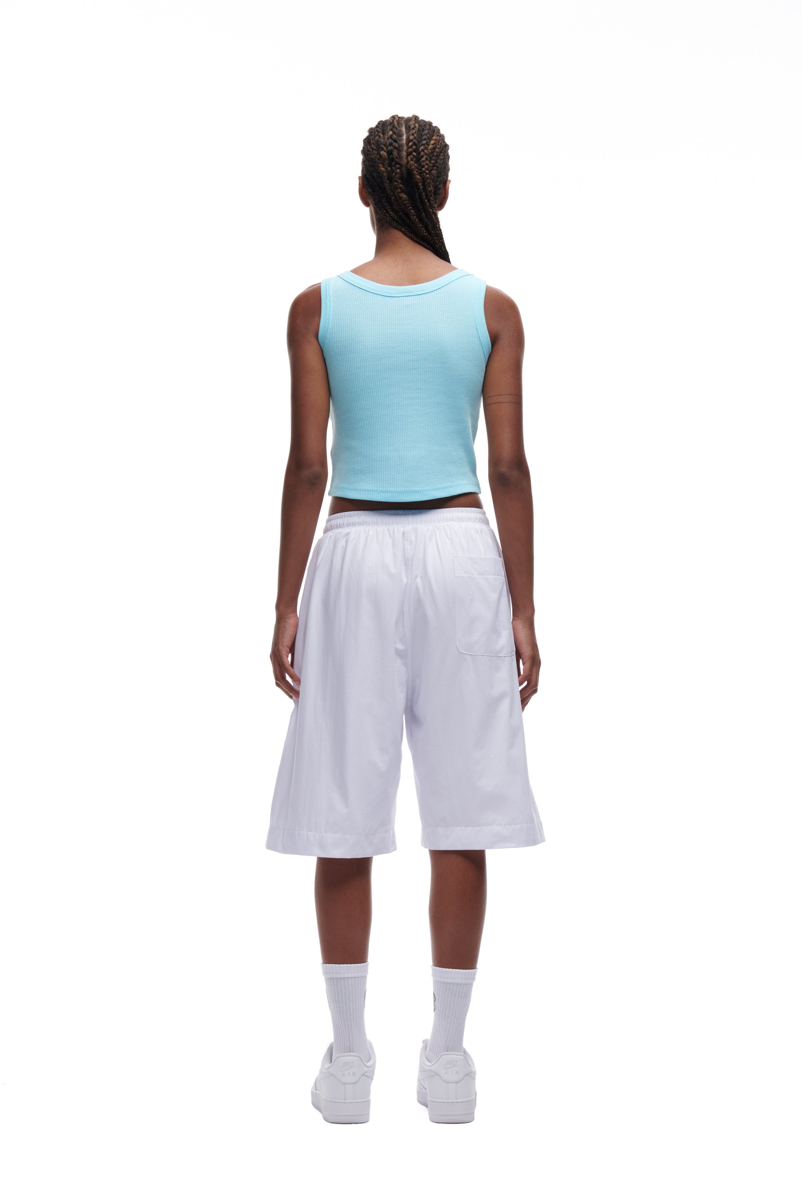 A person with braided hair stands facing away from the camera, wearing a light blue sleeveless top, 6PM BAGGY SHORTS WHITE, white socks, and white sneakers against a white background.