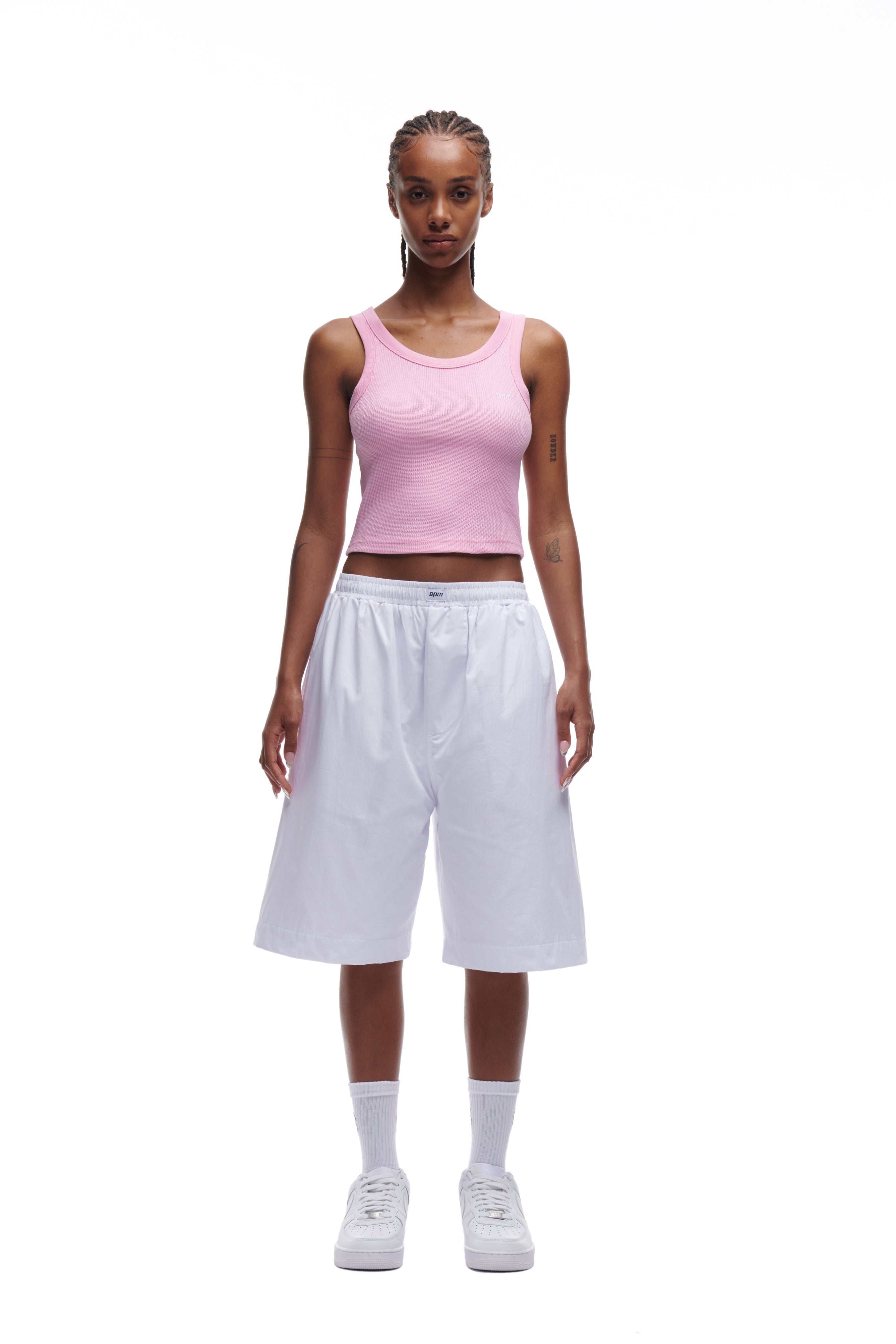 A woman faces forward against a plain white background, wearing the 6PM WAFFLE KNIT CROP TOP BABY PINK, loose white knee-length shorts, white crew socks, and white sneakers.