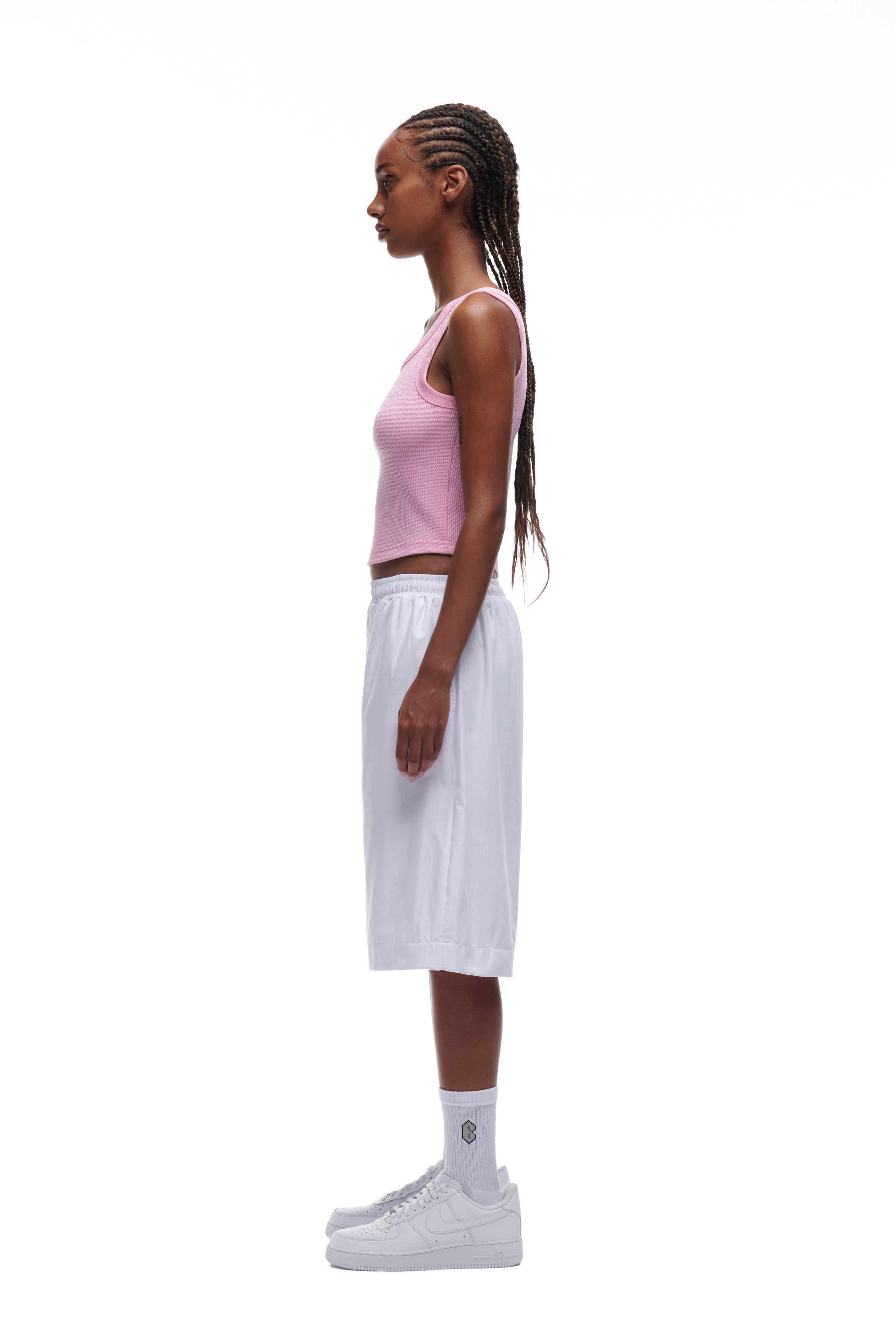 A woman with long braided hair stands in profile against a white background, wearing the 6PM WAFFLE KNIT CROP TOP in baby pink, paired with loose white knee-length shorts, white socks, and white sneakers.