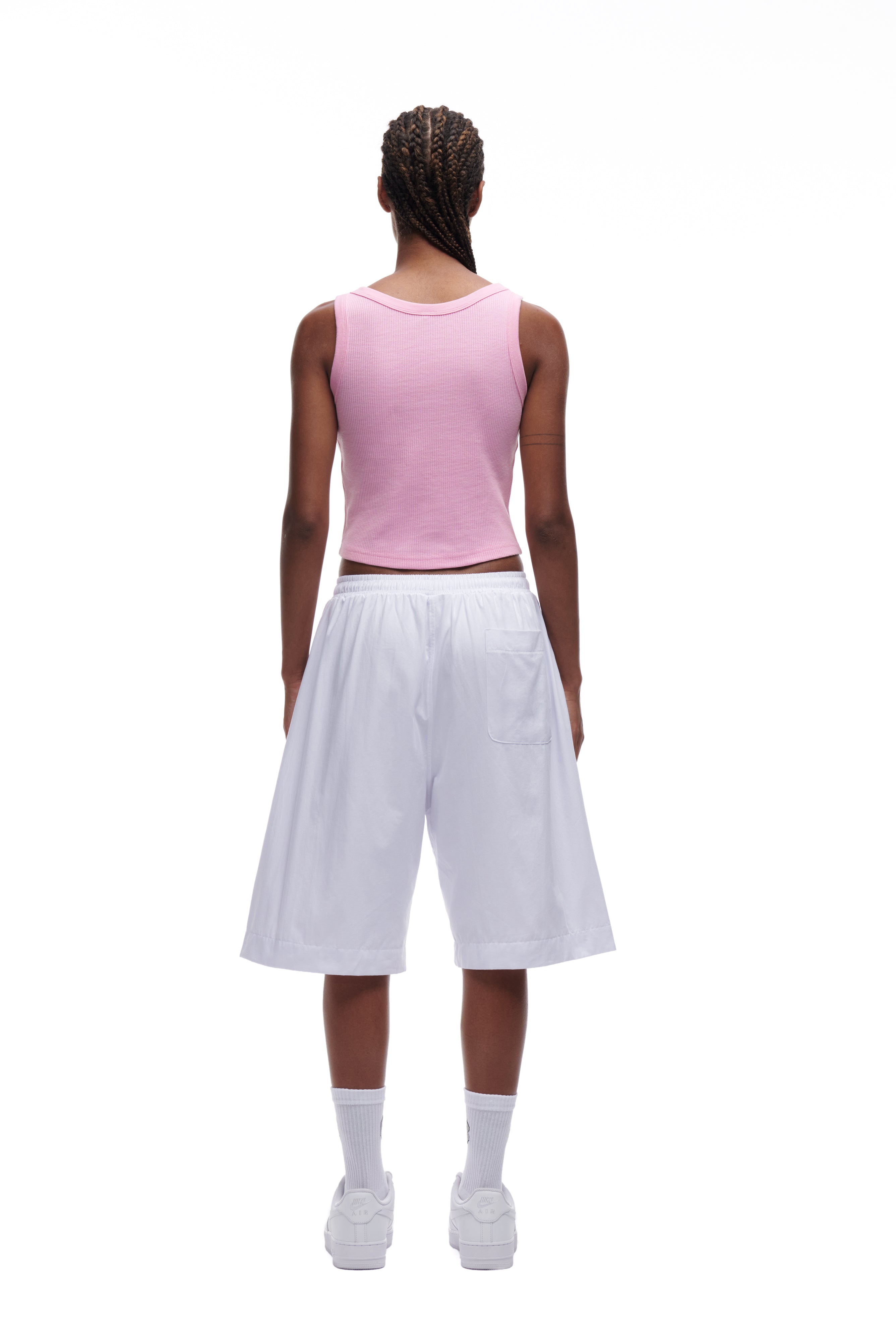A person with braided hair stands against a white background, wearing the 6PM WAFFLE KNIT CROP TOP in baby pink, paired with white knee-length wide shorts, white crew socks, and white sneakers.