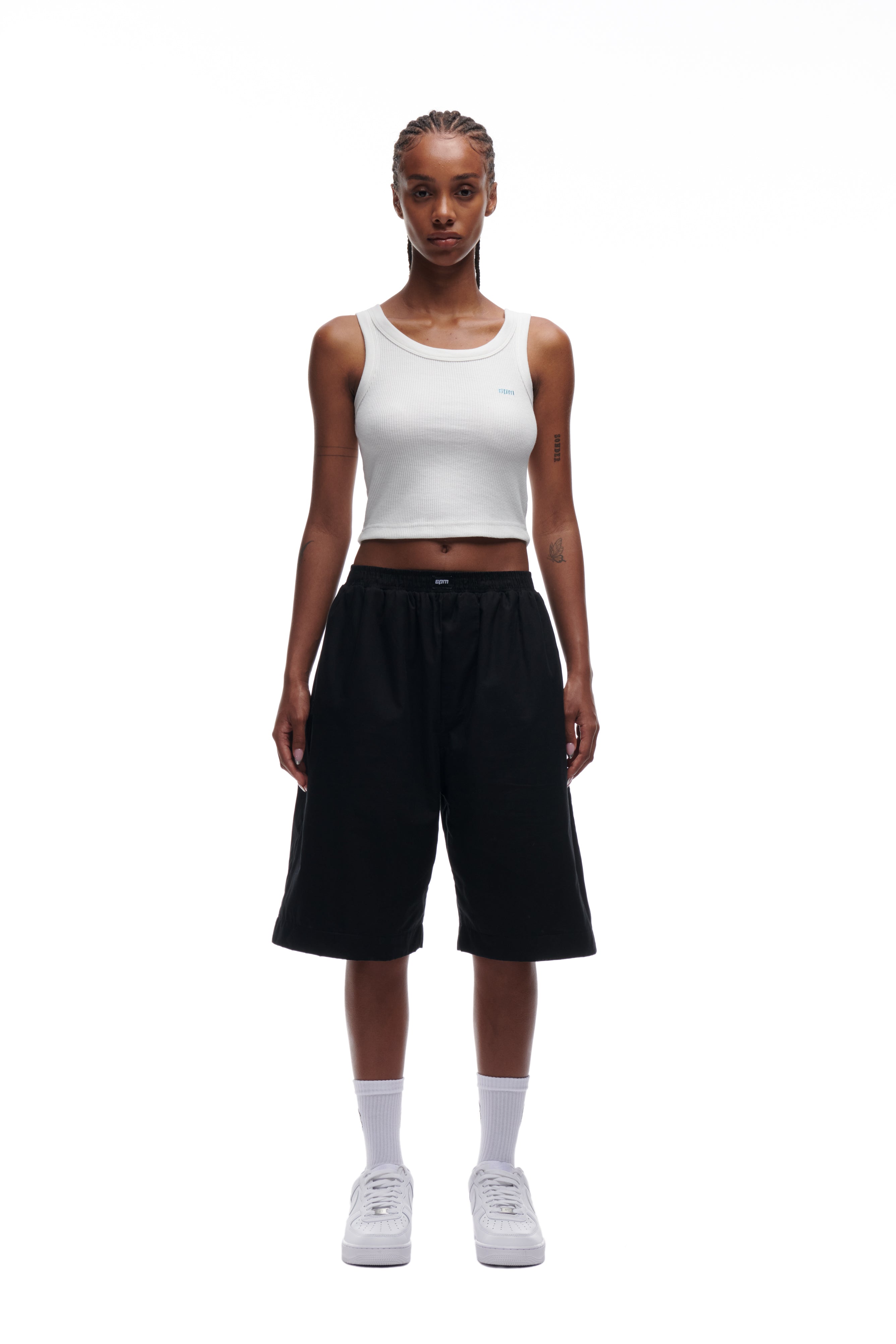 A person stands facing forward, wearing the 6PM WAFFLE KNIT CROP TOP WHITE, loose black knee-length shorts, white crew socks, and white sneakers against a plain white background.
