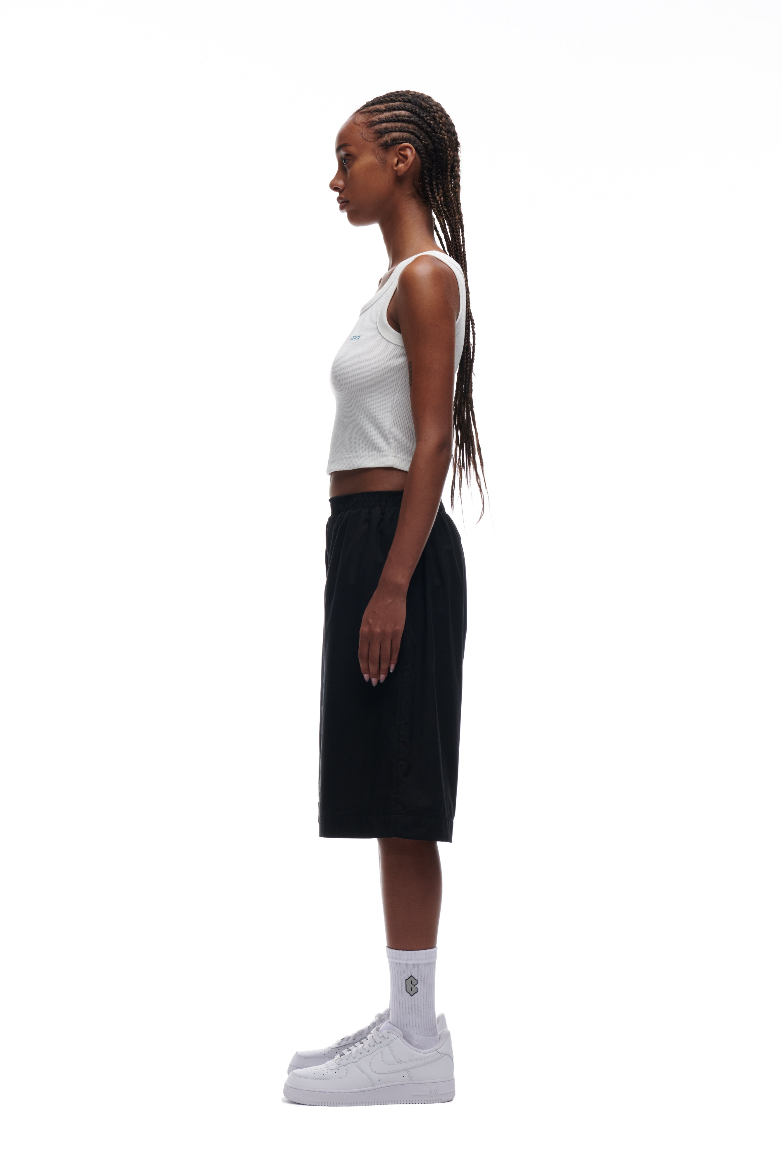 A woman with long braided hair stands in profile against a white background, wearing the 6PM WAFFLE KNIT CROP TOP in white, paired with black knee-length shorts, white crew socks, and white sneakers.