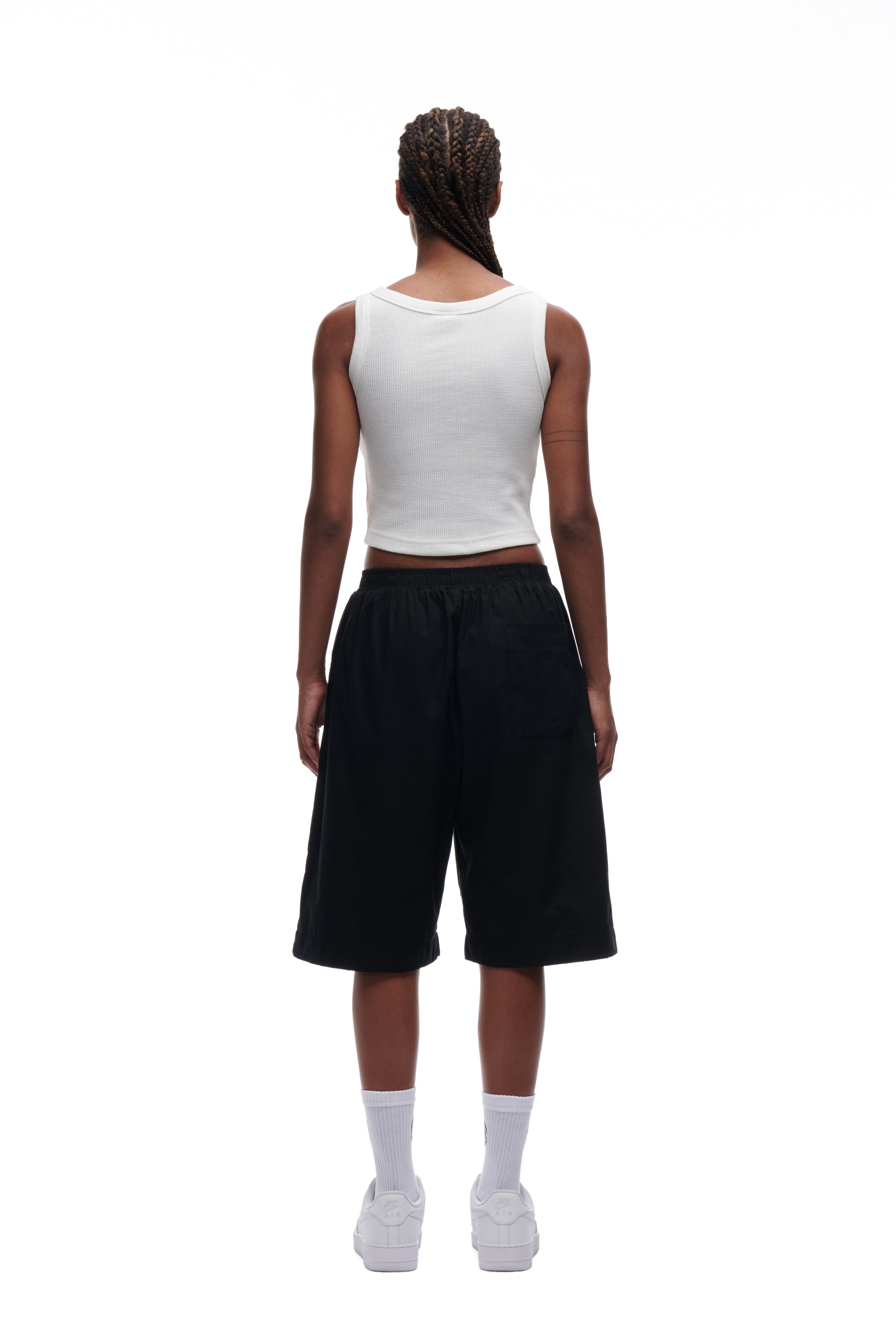 A person with braided hair faces away, wearing the 6PM WAFFLE KNIT CROP TOP in white, paired with loose black shorts, white socks, and white sneakers, set against a plain white background.