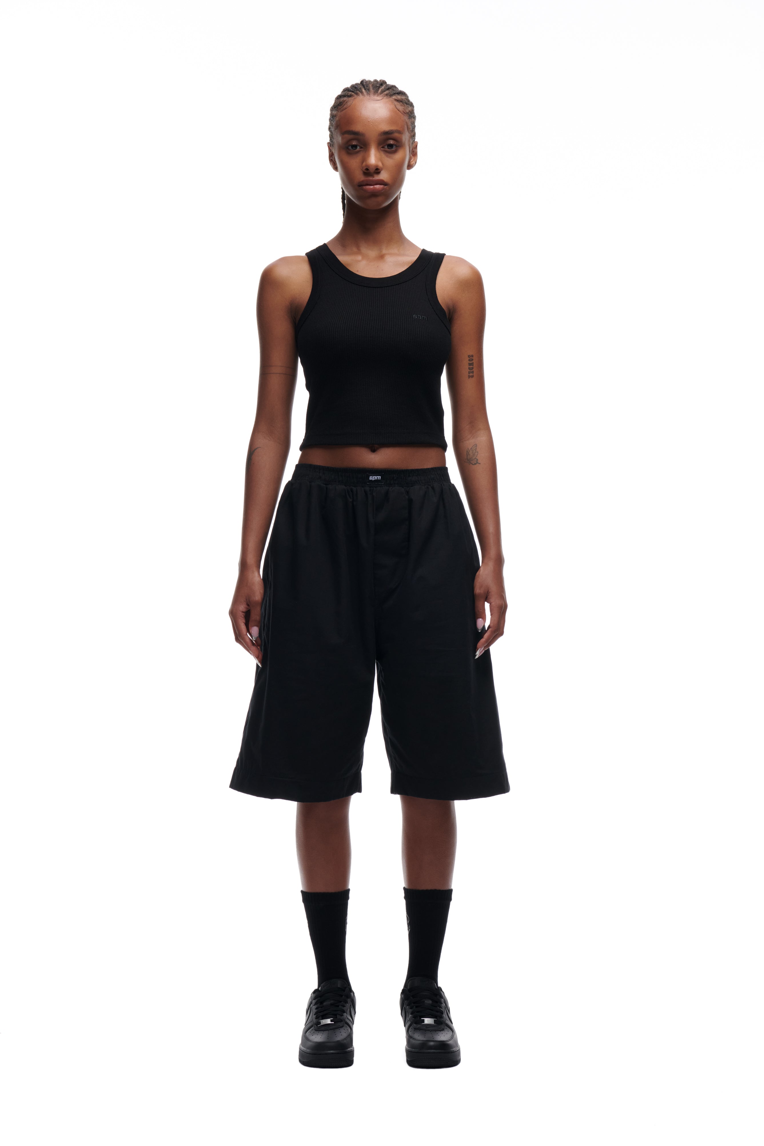 A person stands facing forward against a white background, wearing the 6PM WAFFLE KNIT CROP TOP BLACK, paired with loose black knee-length shorts, black socks, and black shoes.