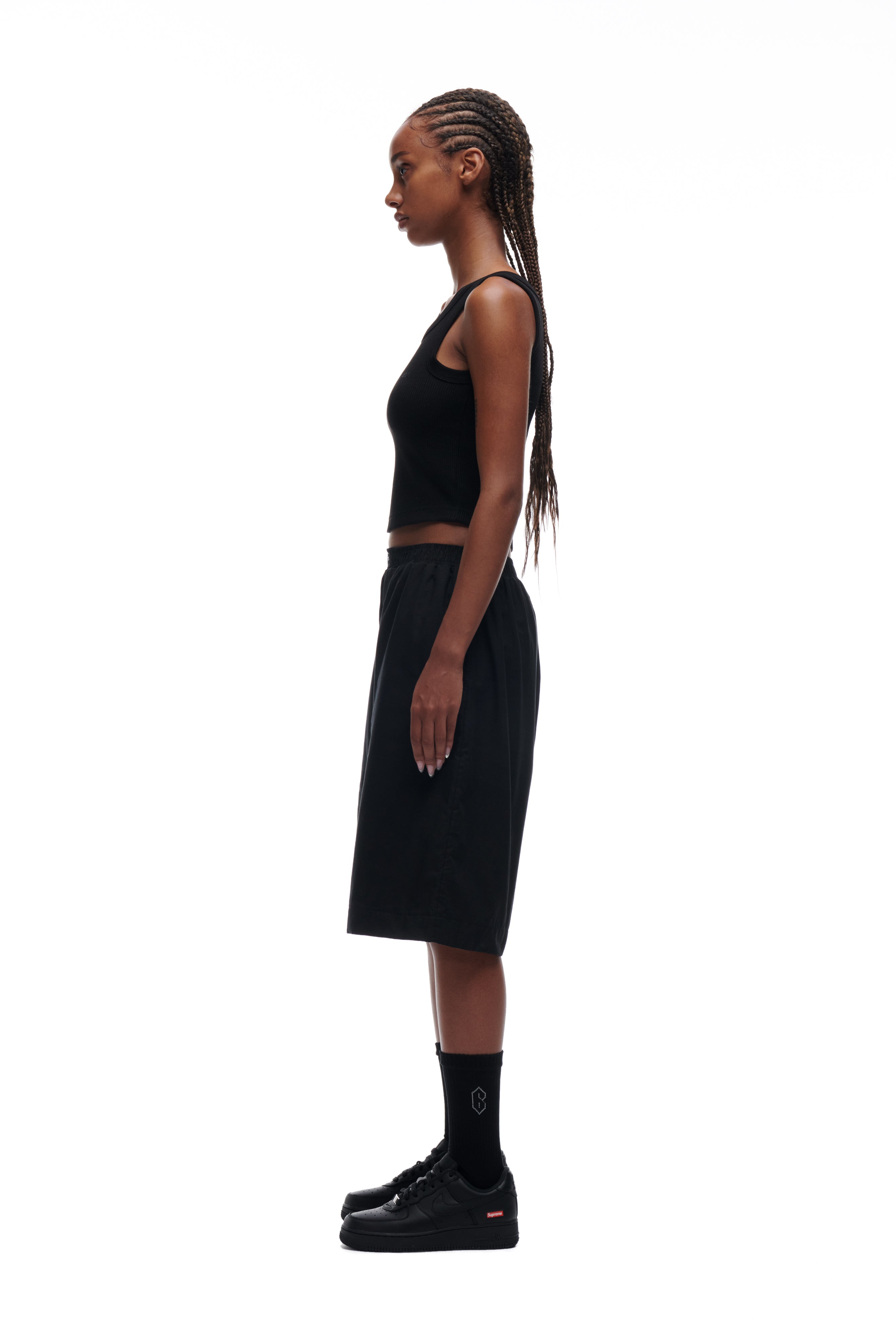 A woman with long braided hair stands in profile against a white background, wearing the 6PM WAFFLE KNIT CROP TOP BLACK with matching black knee-length shorts, black socks, and black sneakers.