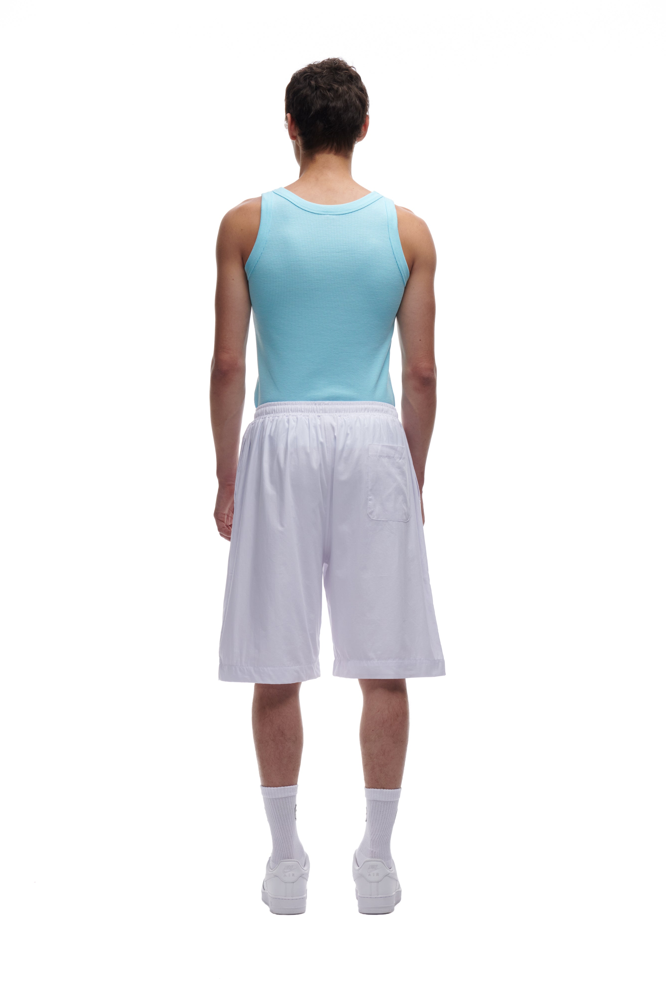 A person with short dark hair wears the 6PM WAFFLE KNIT TANK TOP BABY BLUE, paired with white knee-length shorts, socks, and sneakers, standing with their back to a plain white background.