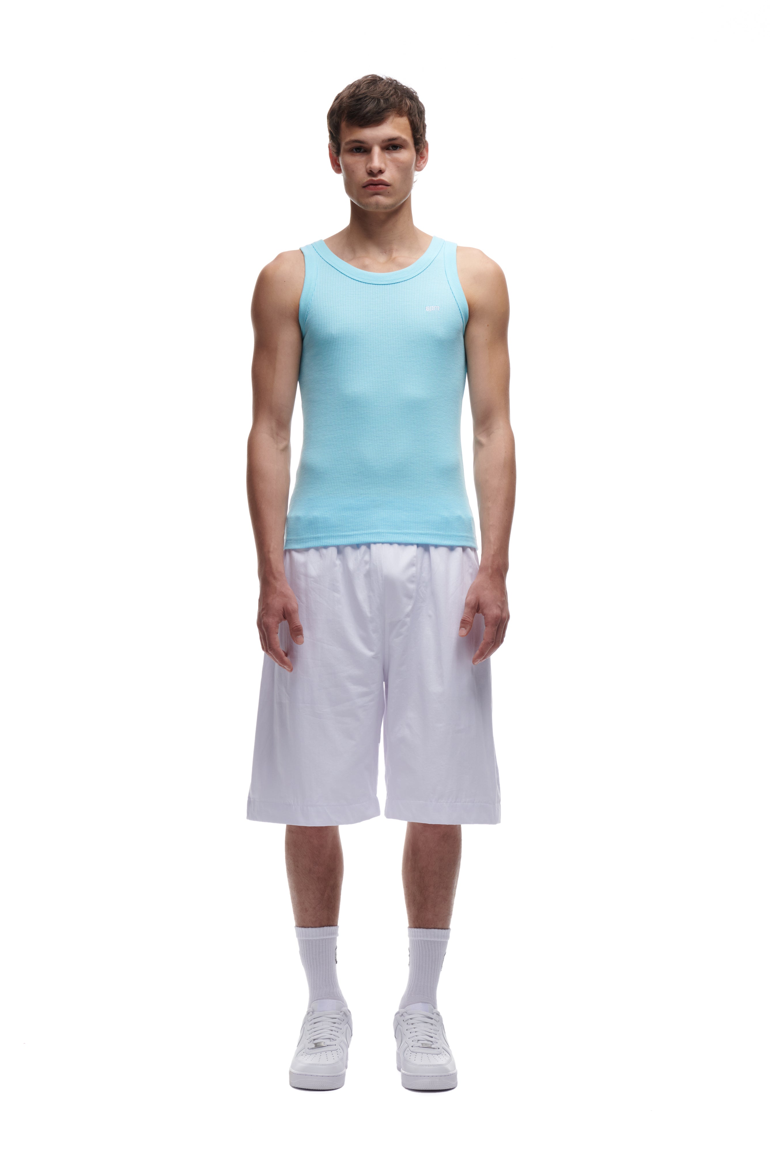 A young man stands facing forward against a white background, wearing the 6PM WAFFLE KNIT TANK TOP BABY BLUE, paired with loose white knee-length shorts, white socks, and white sneakers.