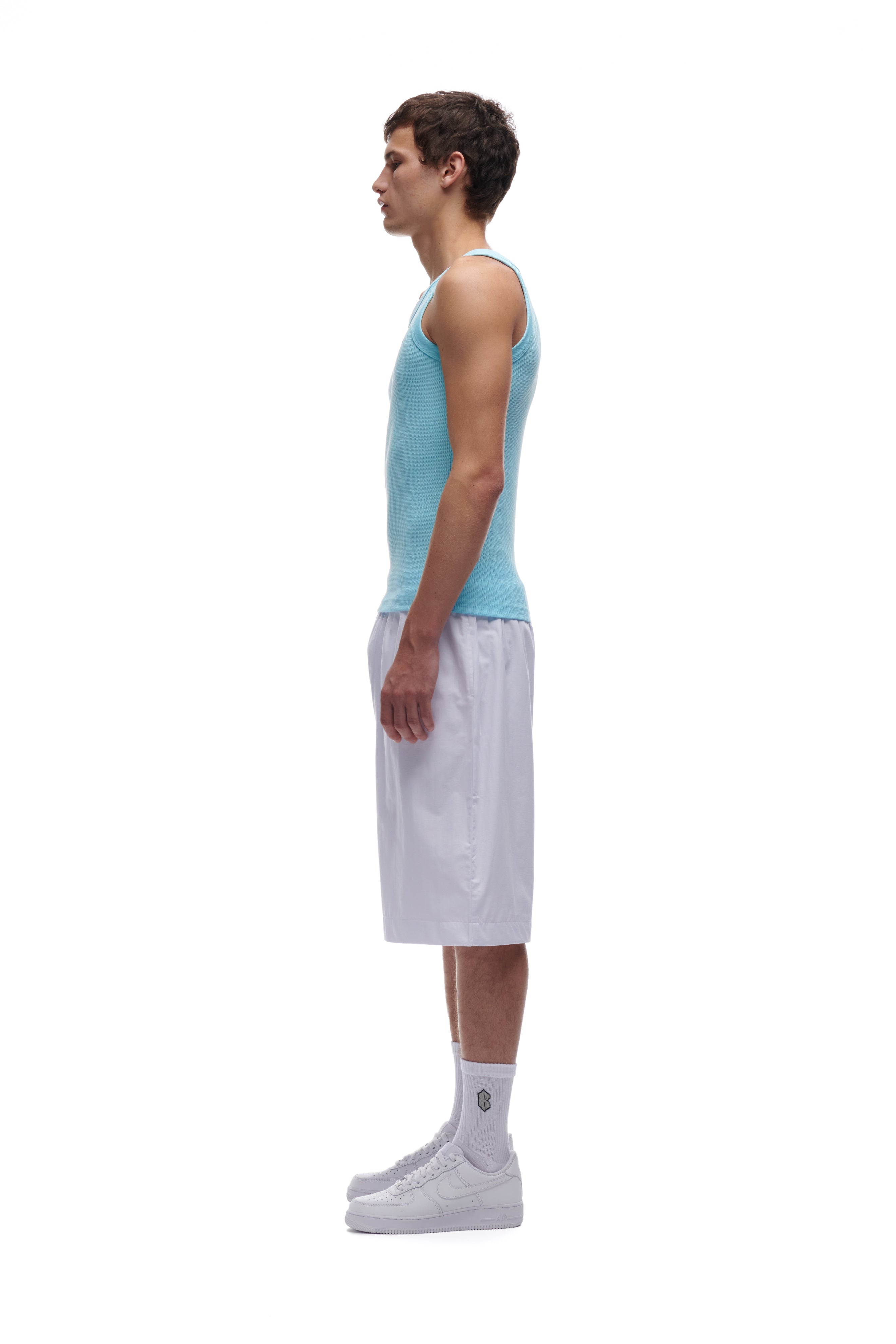 A young man stands in profile against a white background, wearing 6PM’s BAGGY SHORTS WHITE along with a light blue tank top, white socks, and white sneakers.