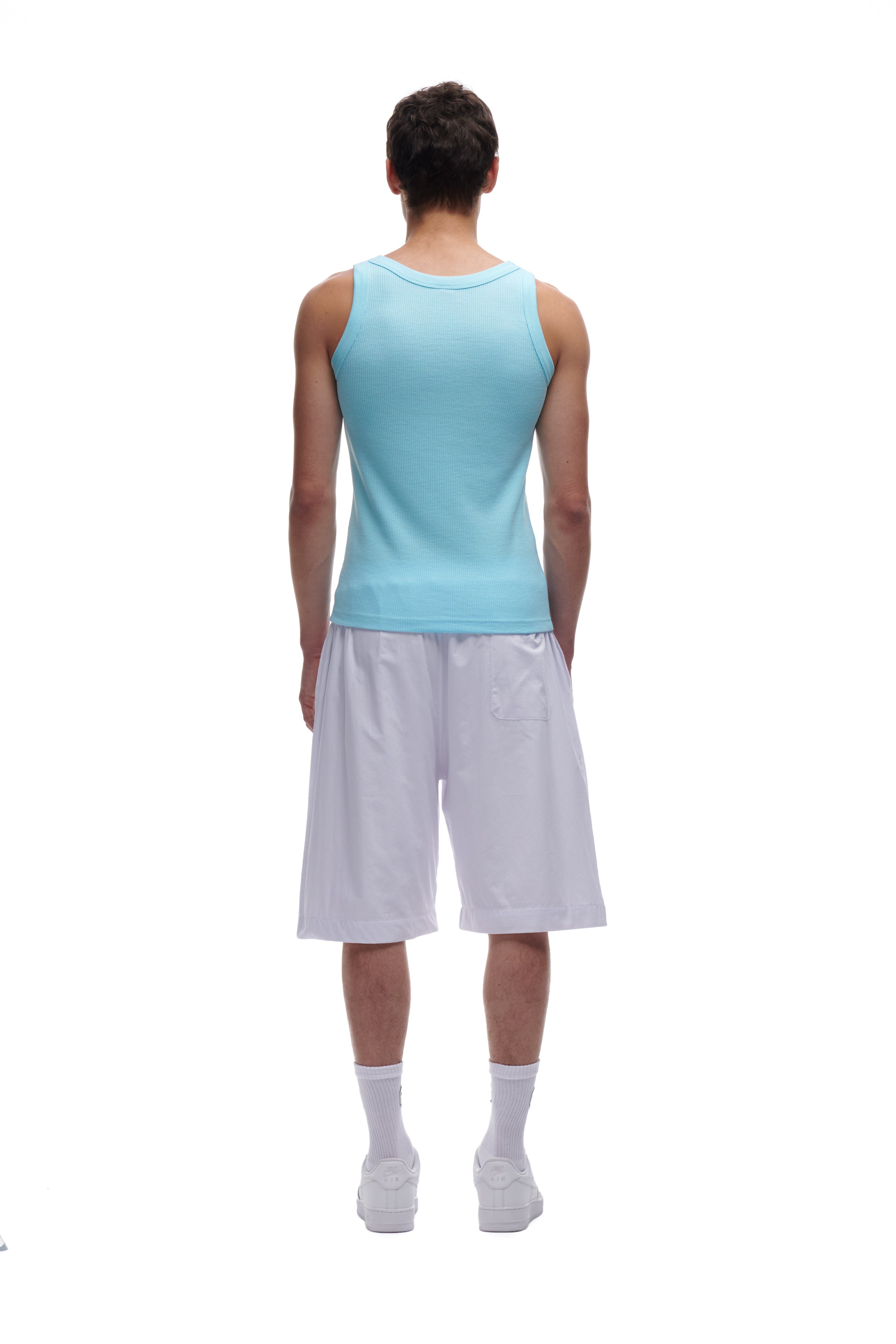 A man stands facing away from the camera, wearing a light blue tank top, 6PM BAGGY SHORTS WHITE, white socks, and white sneakers against a plain white background.