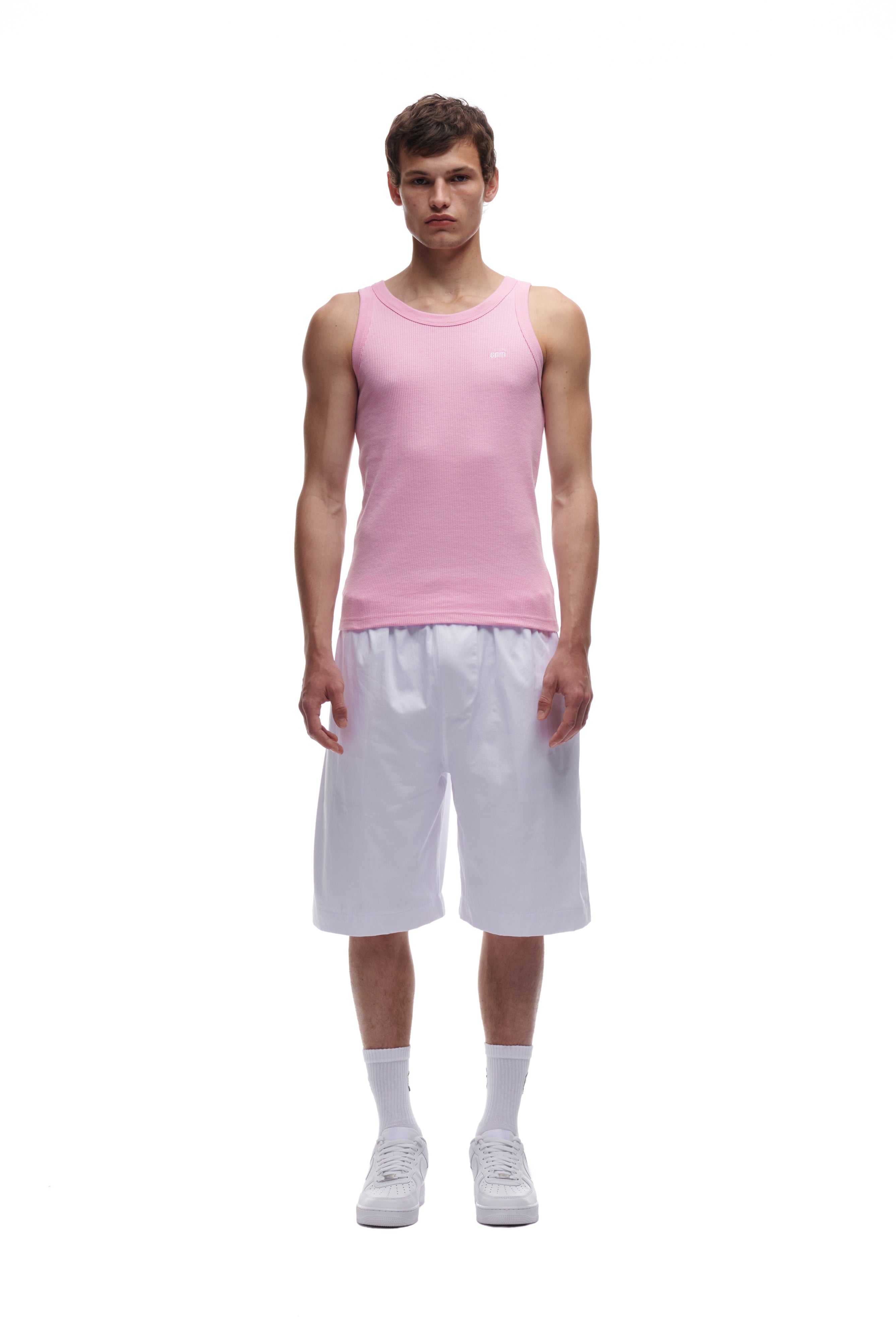 A young man faces forward against a white background, wearing the 6PM WAFFLE KNIT TANK TOP BABY PINK, loose white shorts, white socks, and white sneakers.