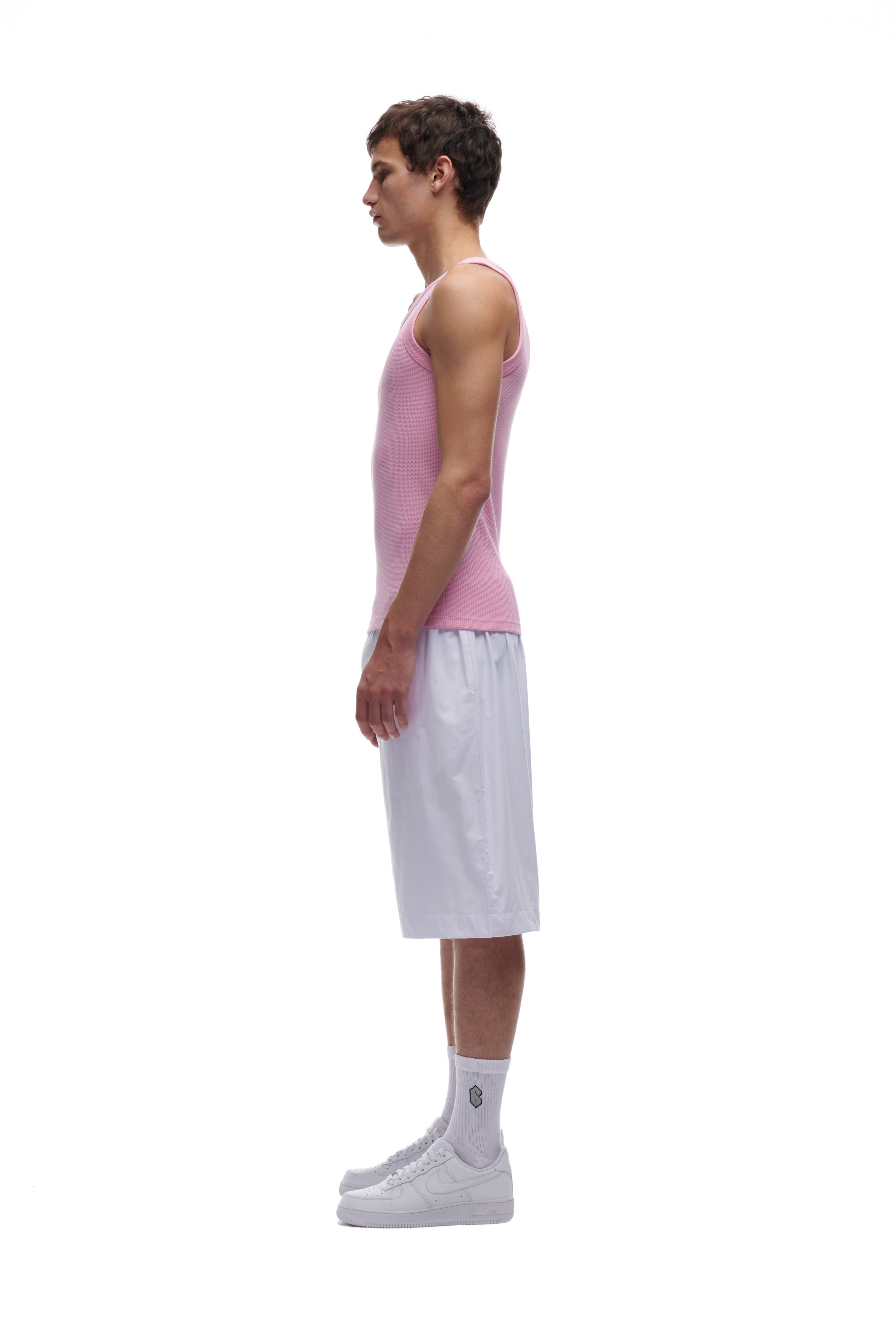 A man in profile against a white background wears the 6PM WAFFLE KNIT TANK TOP BABY PINK, paired with white knee-length shorts, crew socks, and sneakers.