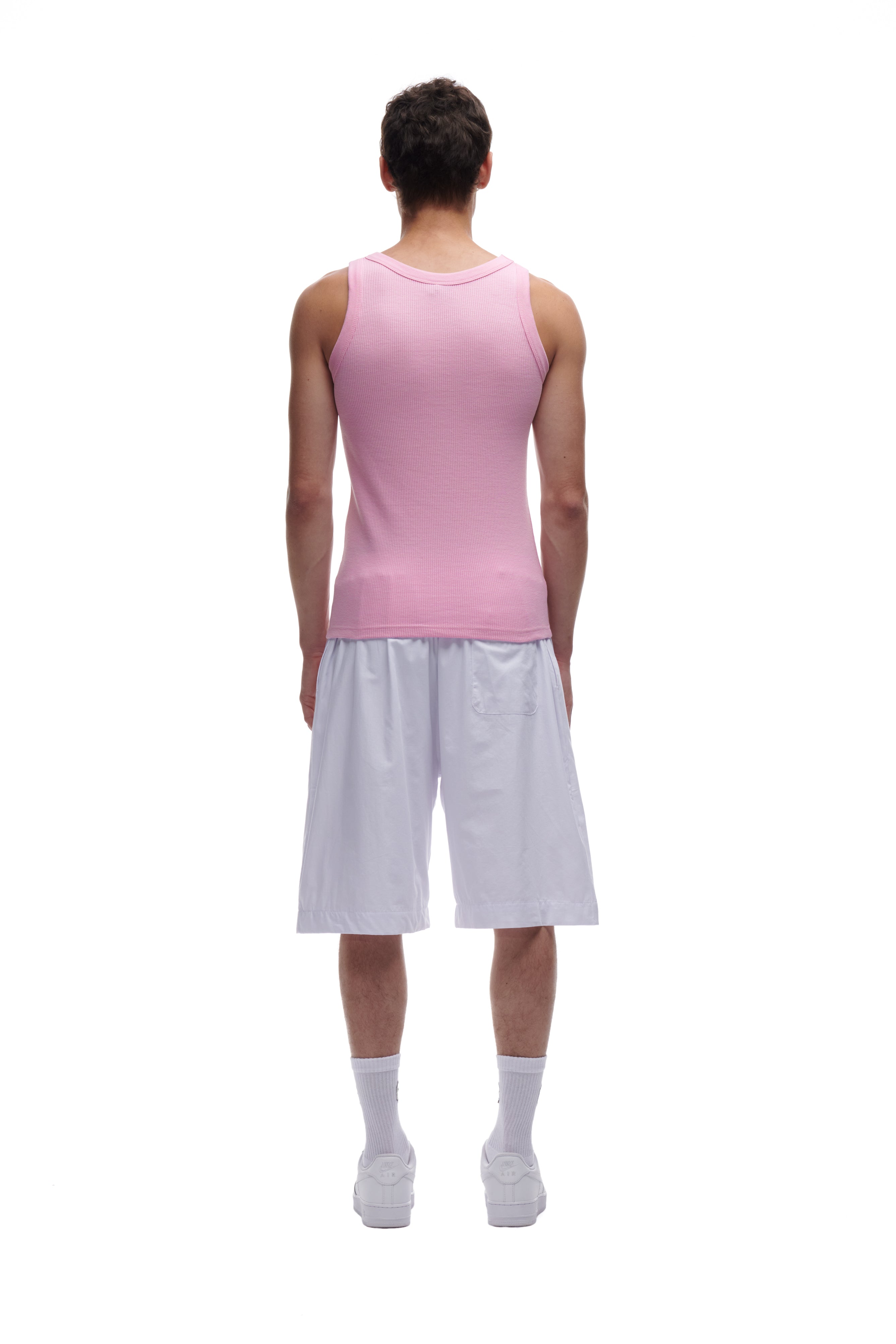 A person faces away against a plain white background, wearing a 6PM WAFFLE KNIT TANK TOP in baby pink, paired with white knee-length shorts, white crew socks, and white sneakers.
