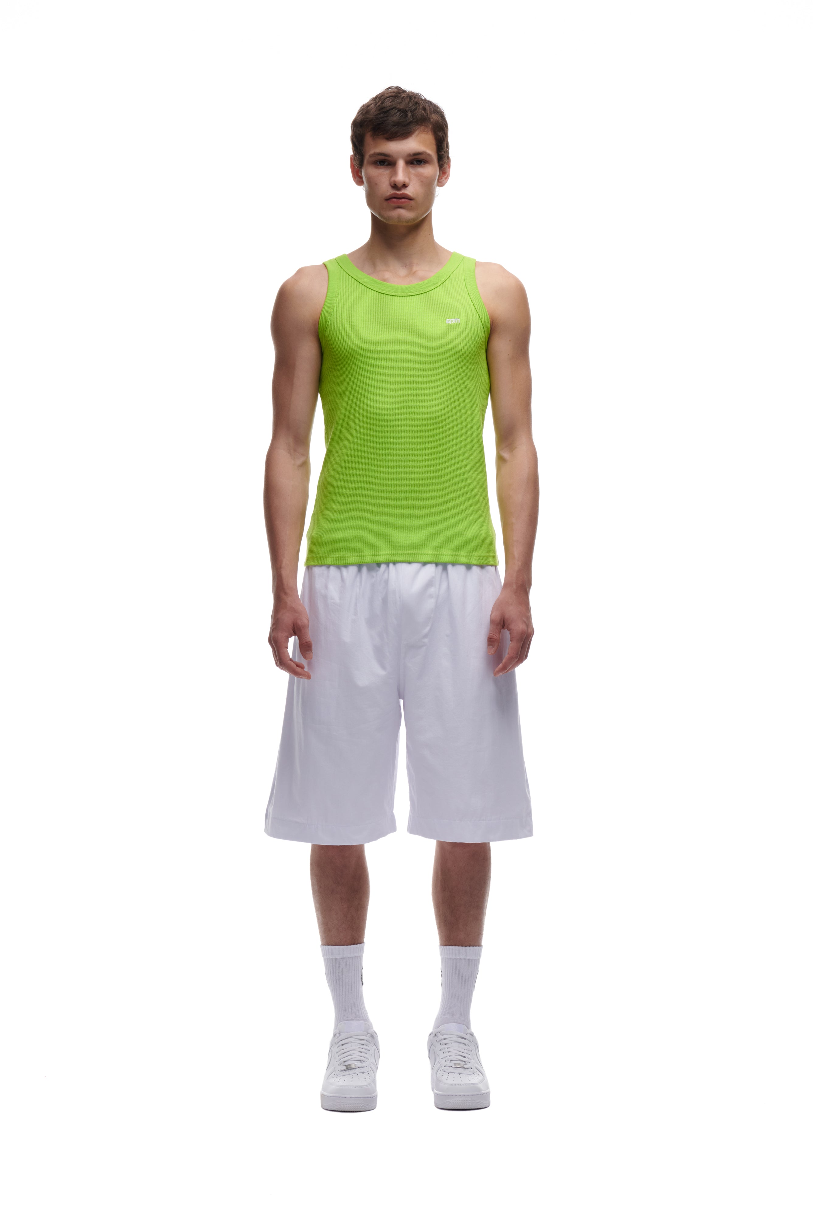 A young man stands facing forward wearing the 6PM WAFFLE KNIT TANK TOP LIME GREEN, paired with loose white knee-length shorts, white socks, and white sneakers, set against a plain white background.