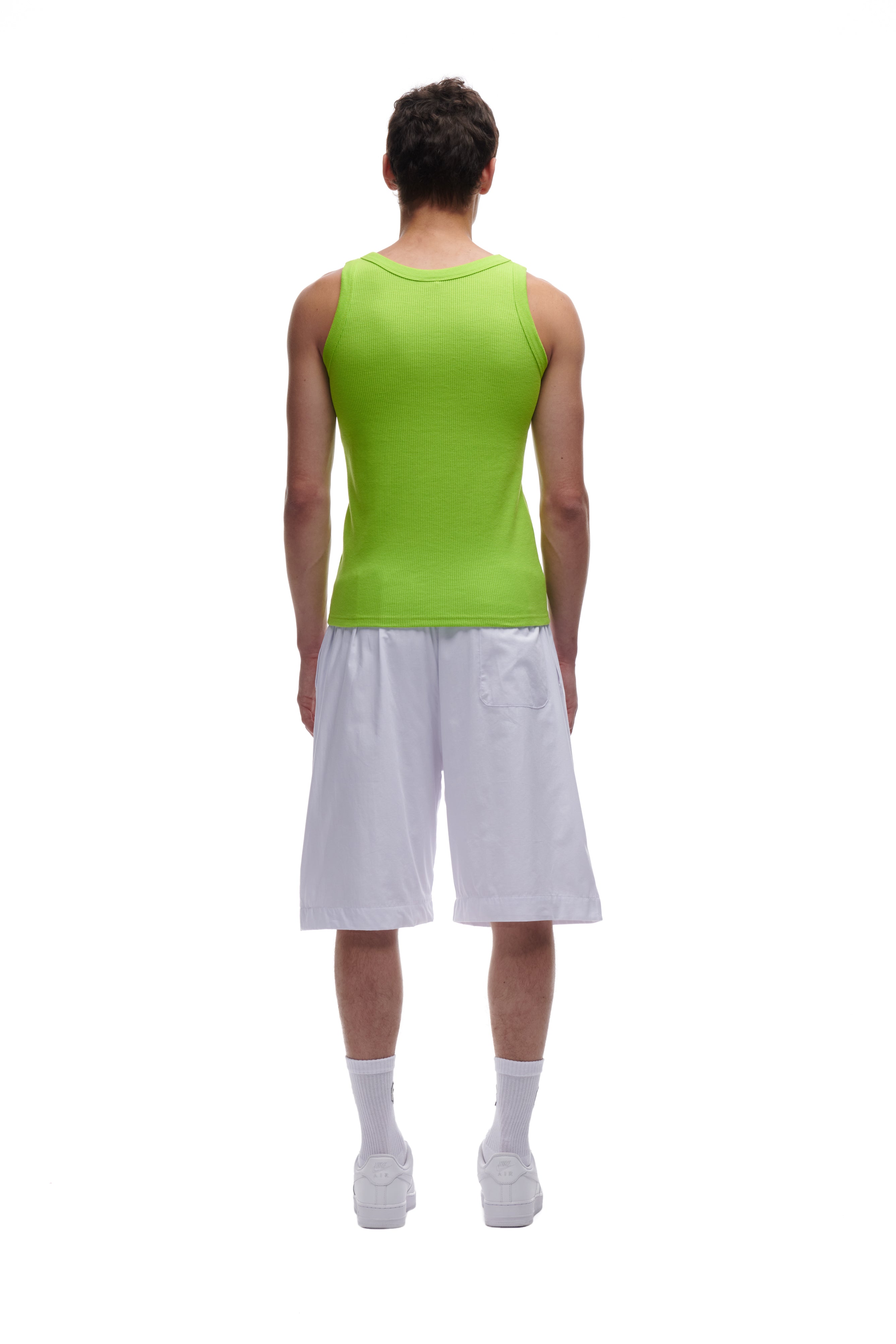 A person stands facing away against a white background, wearing the 6PM WAFFLE KNIT TANK TOP LIME GREEN, loose white knee-length shorts, white socks, and white sneakers.