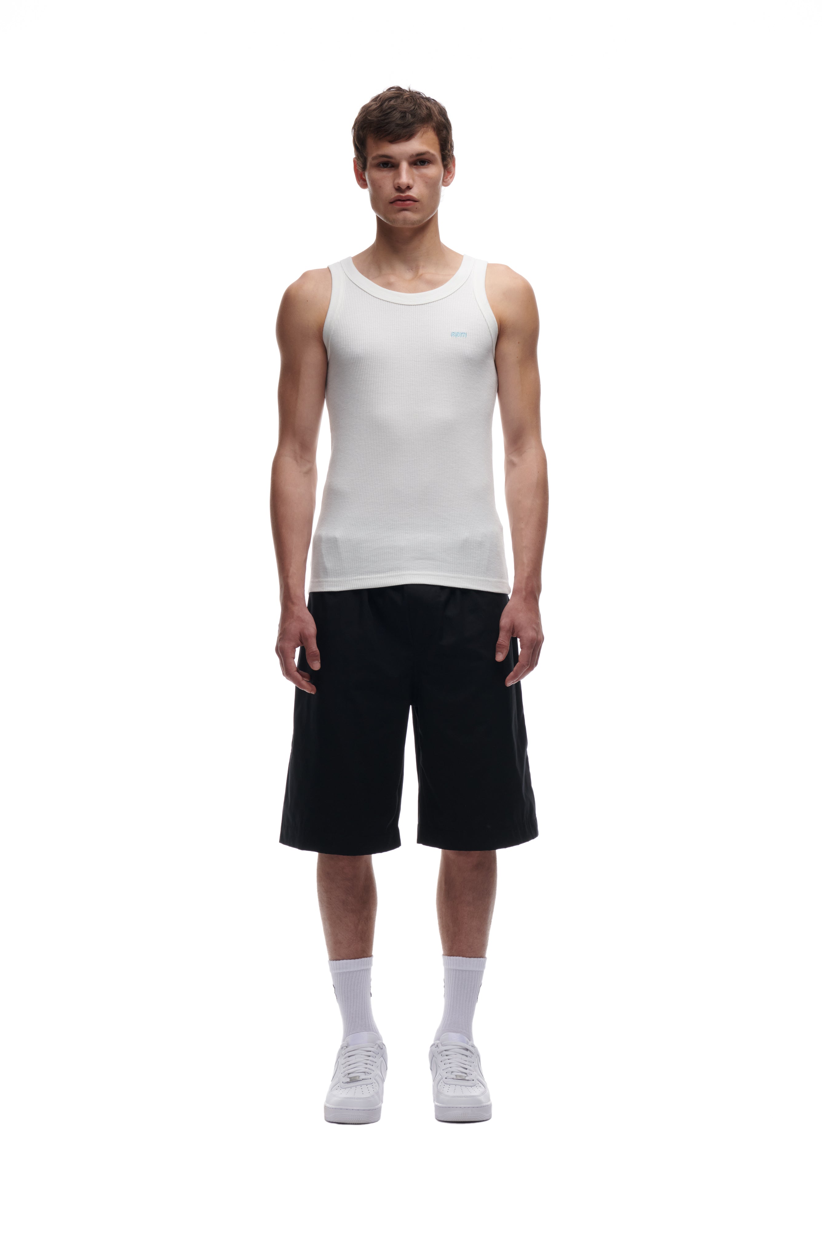 A young man faces forward against a plain white background, wearing the 6PM WAFFLE KNIT TANK TOP WHITE, loose black knee-length shorts, white crew socks, and white sneakers.