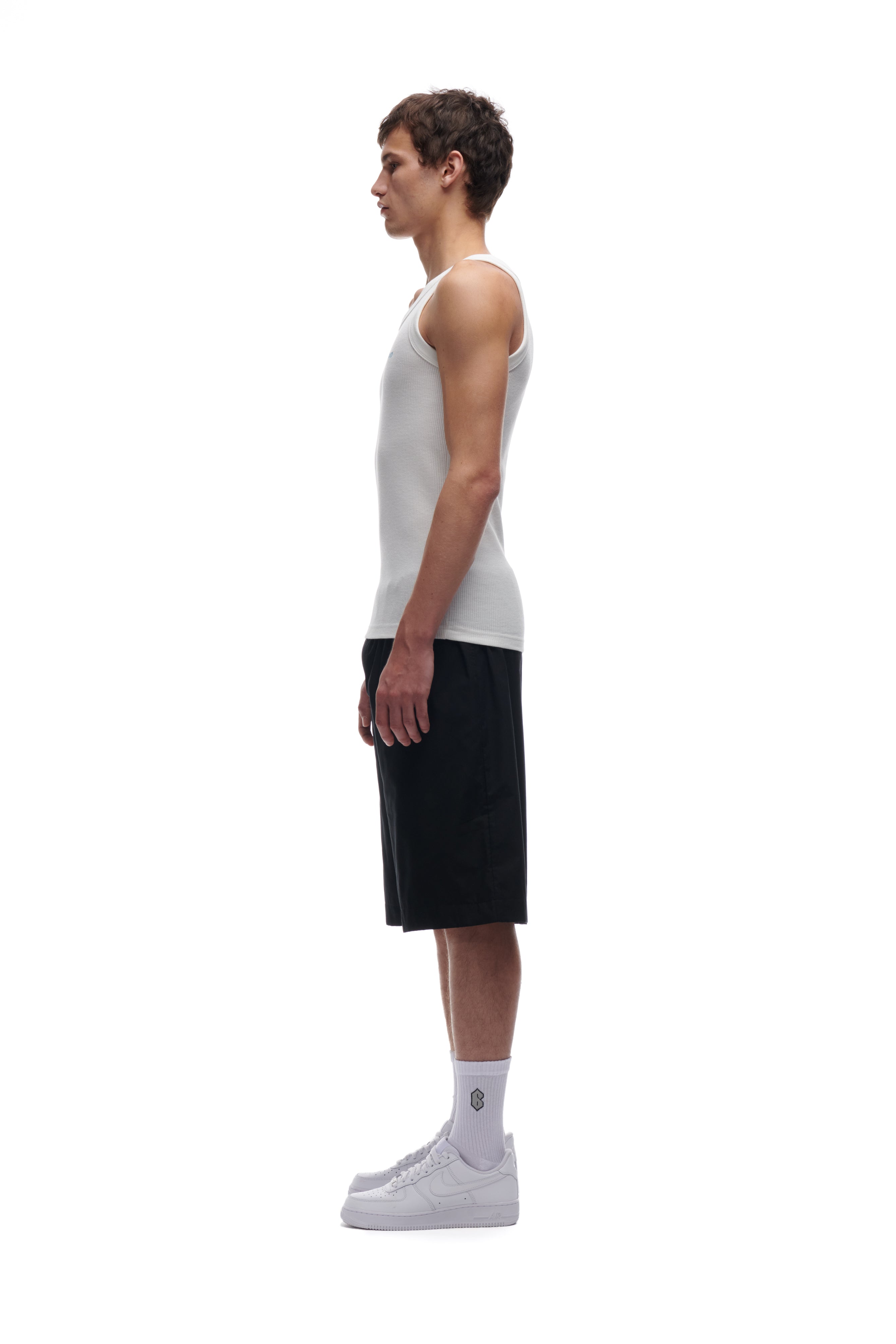 A young man stands in profile against a white background, wearing the 6PM WAFFLE KNIT TANK TOP WHITE, paired with black shorts, white socks, and white sneakers. His arms rest naturally at his sides.
