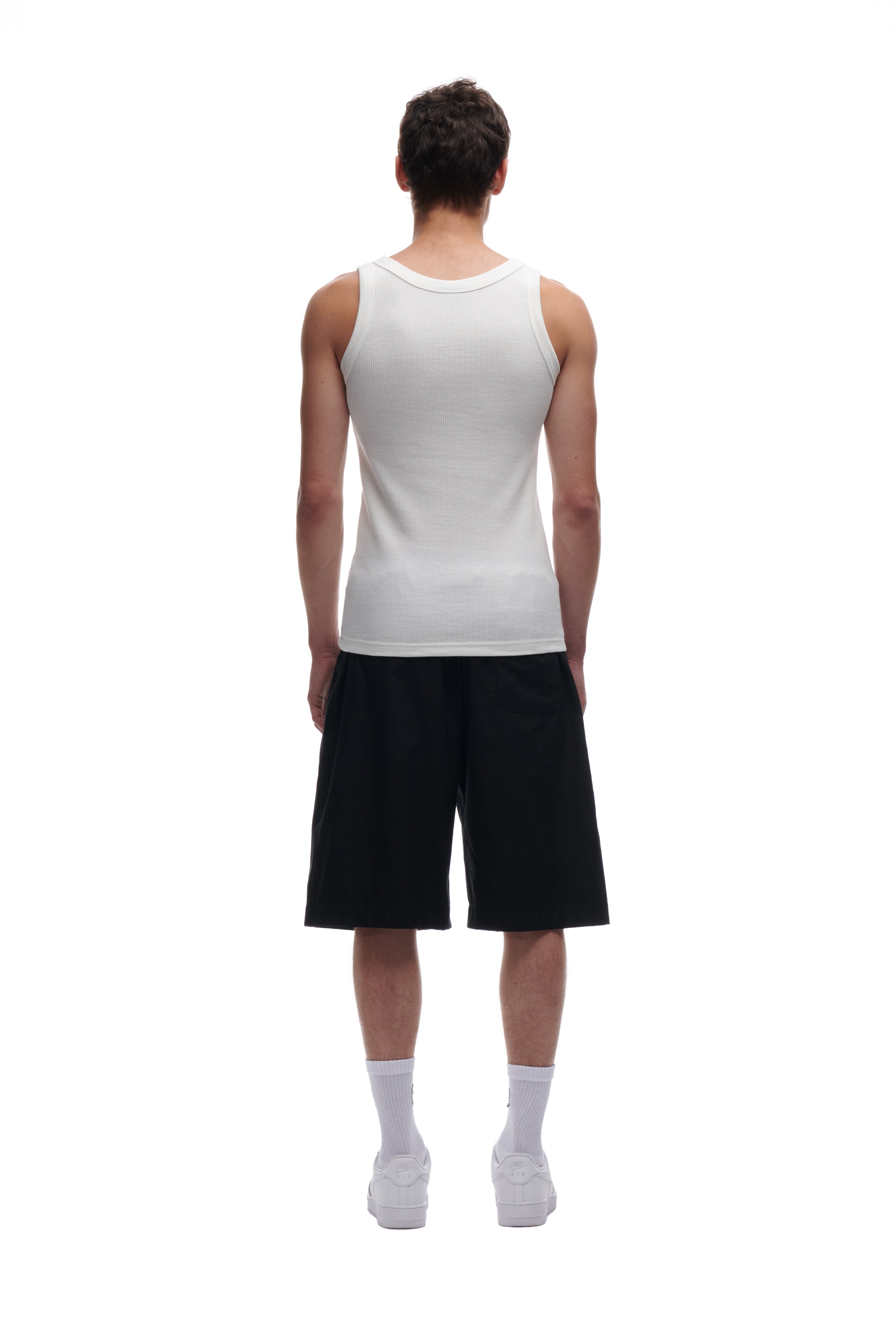 A man faces away from the camera against a white background, wearing the 6PM WAFFLE KNIT TANK TOP WHITE, black knee-length shorts, white socks, and white sneakers.