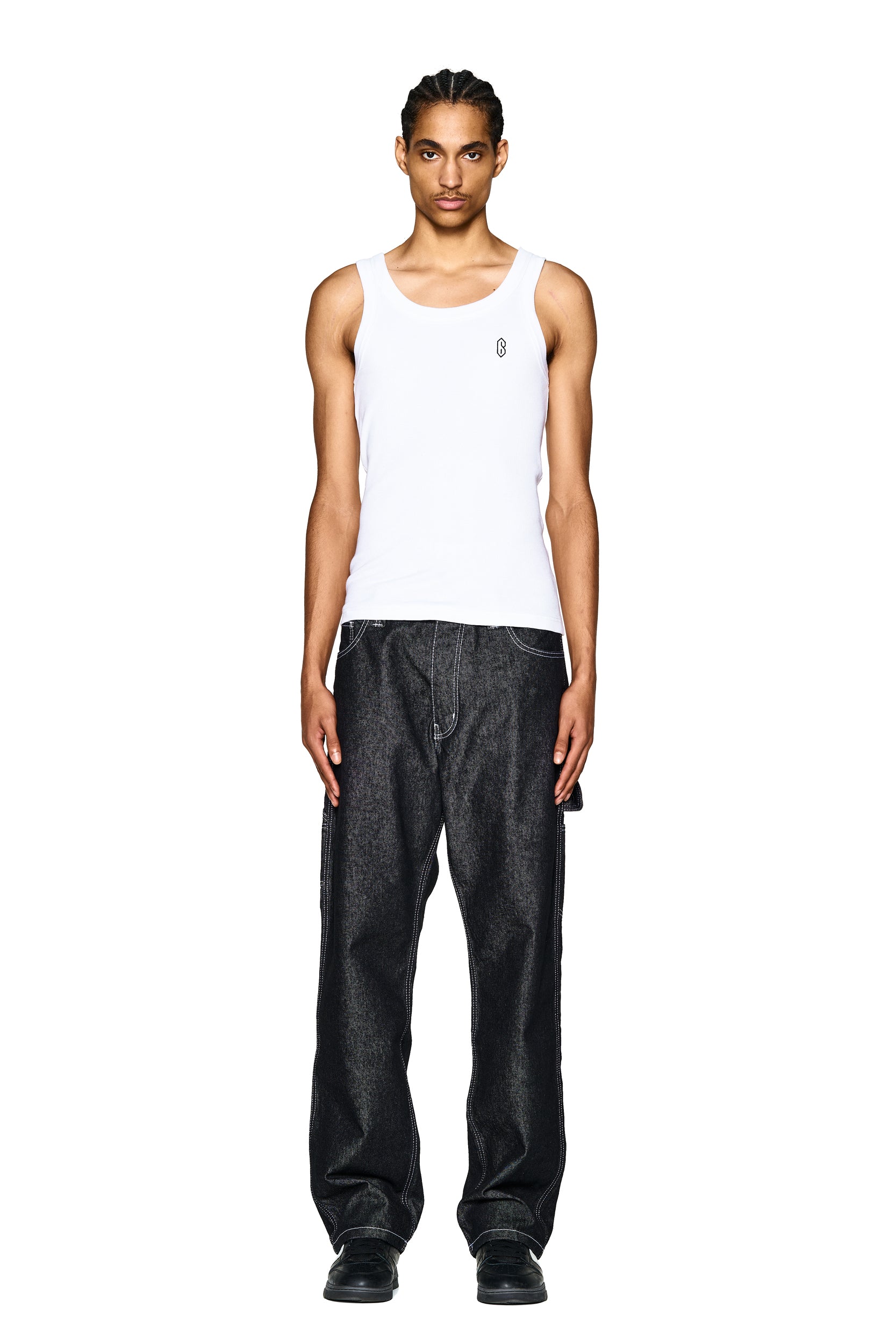 A person with braided hair, facing forward against a plain white background, wears 6PM's LOGO DENIM JEANS BLACK paired with a white tank top featuring a small logo and black shoes.