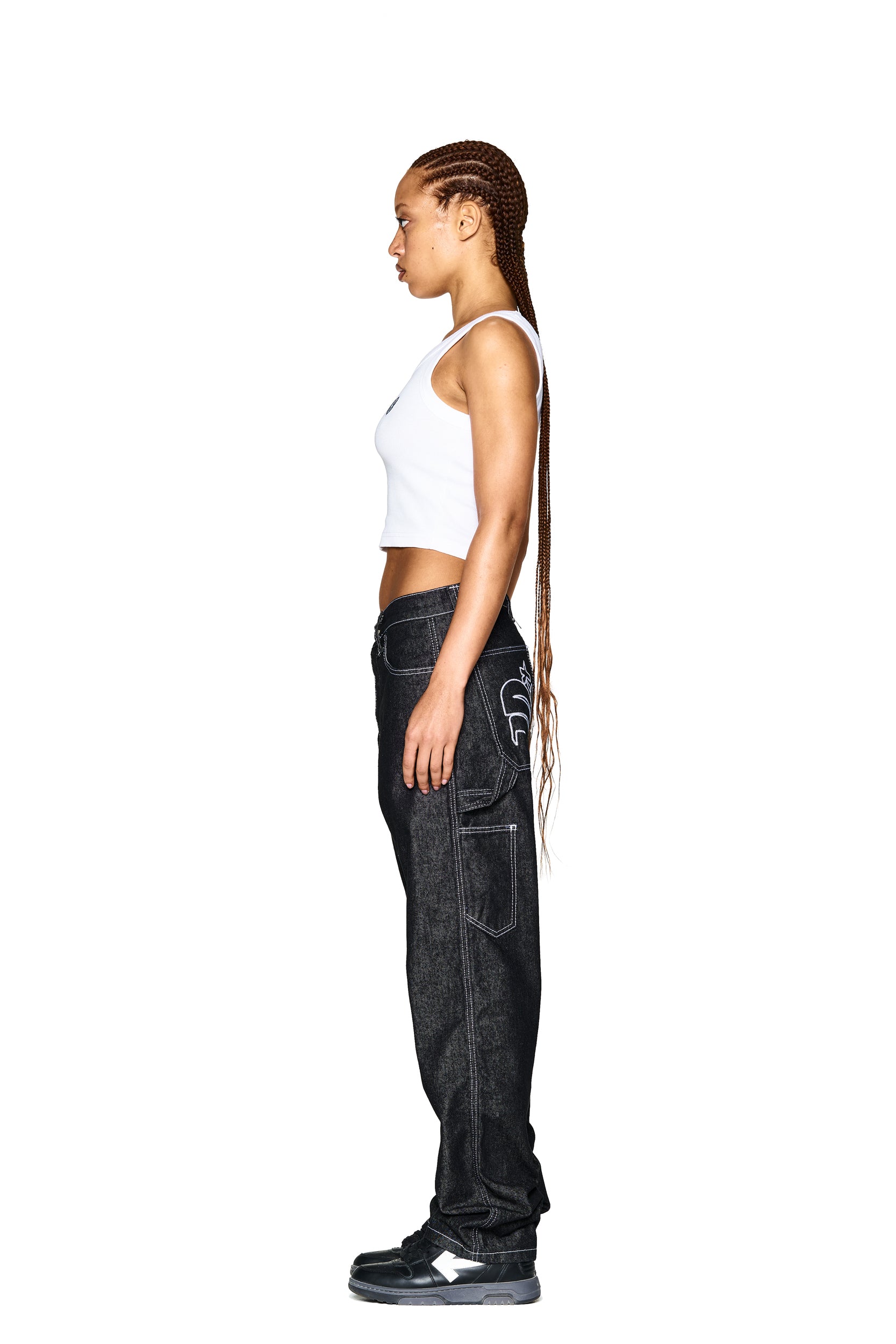 A woman with long braided hair stands in profile, modeling 6PM’s LOGO DENIM JEANS BLACK with visible stitching, paired with a white tank top and black sneakers, against a plain white background.