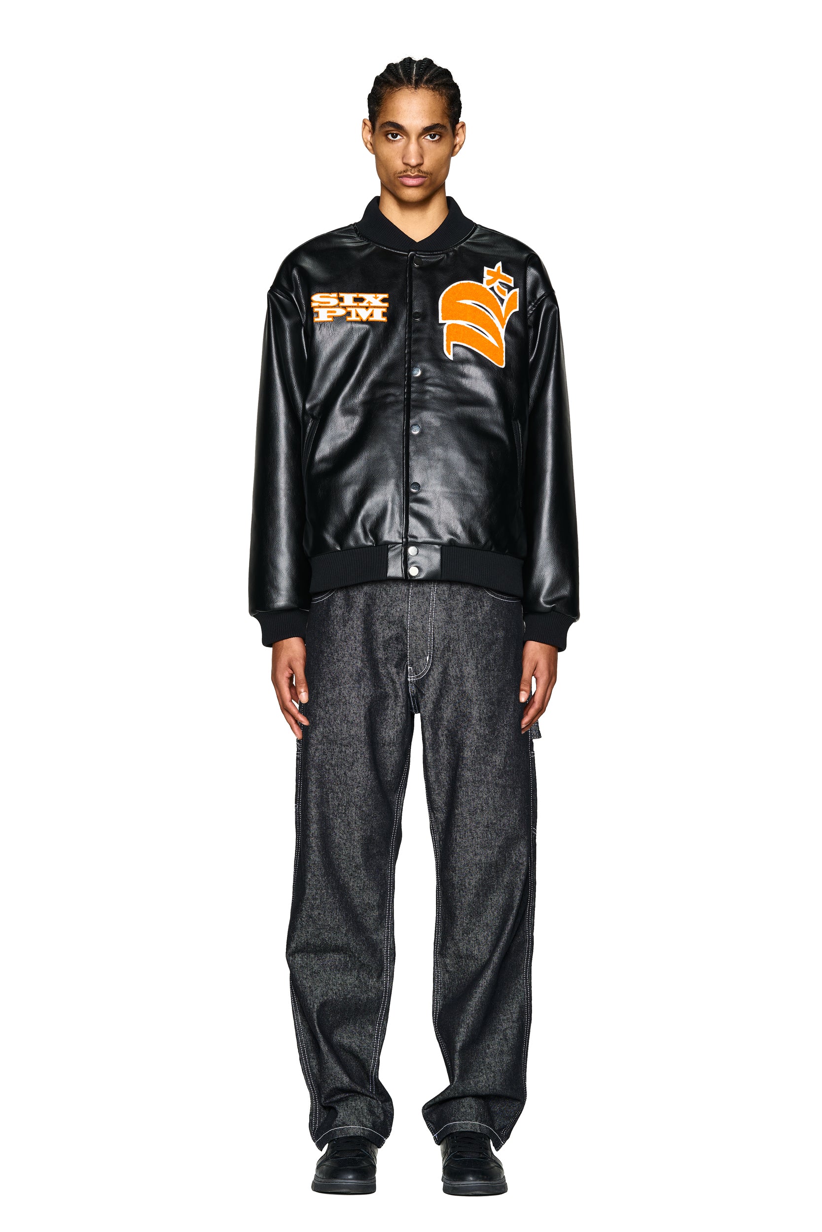 A person with braided hair faces forward against a white background, wearing the 6PM COLLEGE JACKET SLAWN BLACK with orange and white patches, paired with loose dark jeans and black shoes.