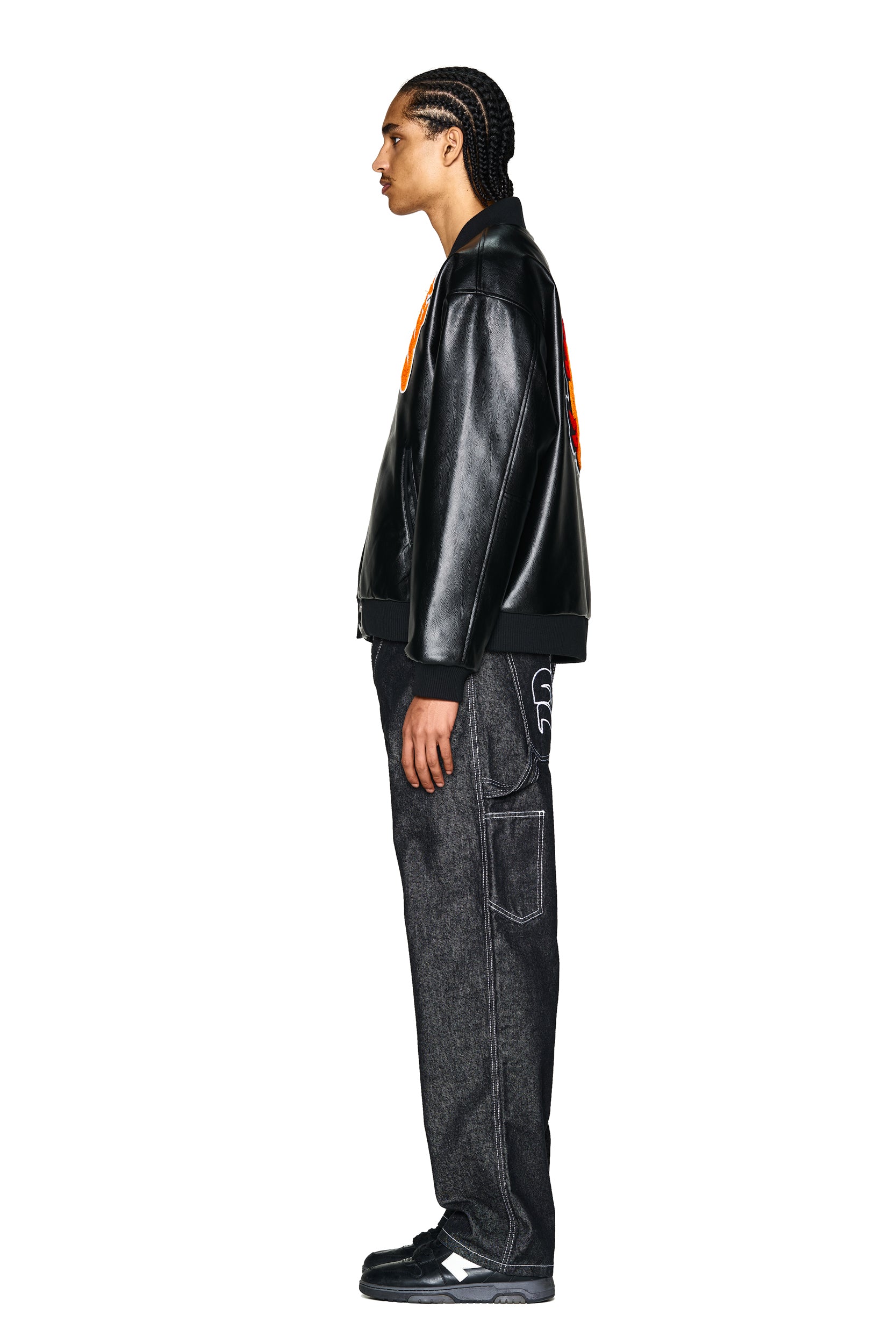 A man with braided hair stands in profile against a plain white background, wearing the 6PM COLLEGE JACKET SLAWN BLACK, paired with dark loose-fitting jeans with white stitching and black sneakers.