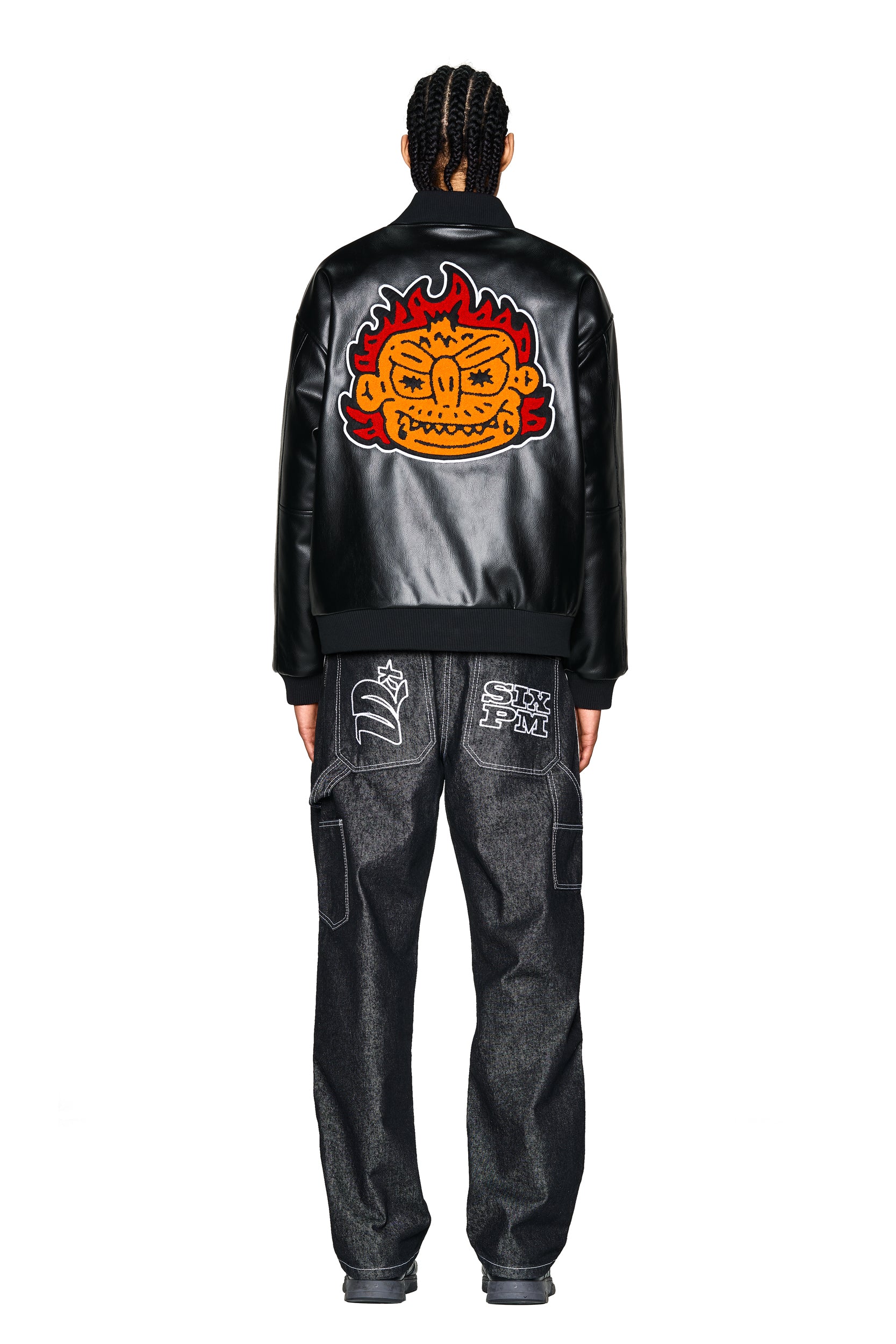 A person with braided hair faces away, wearing the 6PM COLLEGE JACKET SLAWN BLACK featuring a bold, colorful cartoon face with flames on the back, paired with dark jeans with white graphic patches on the back pockets.