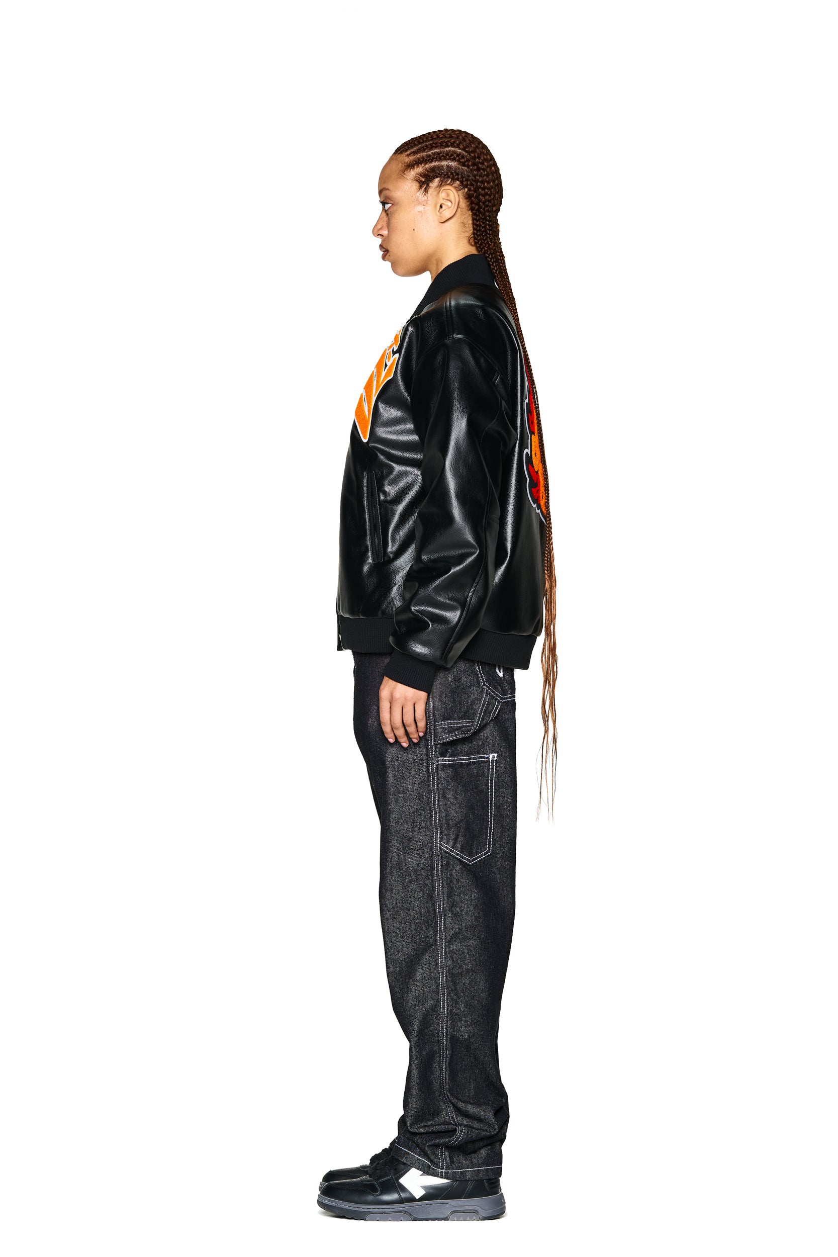 A person with long braided hair stands in profile against a white background, wearing the 6PM COLLEGE JACKET SLAWN BLACK, dark jeans with white stitching, and black sneakers.