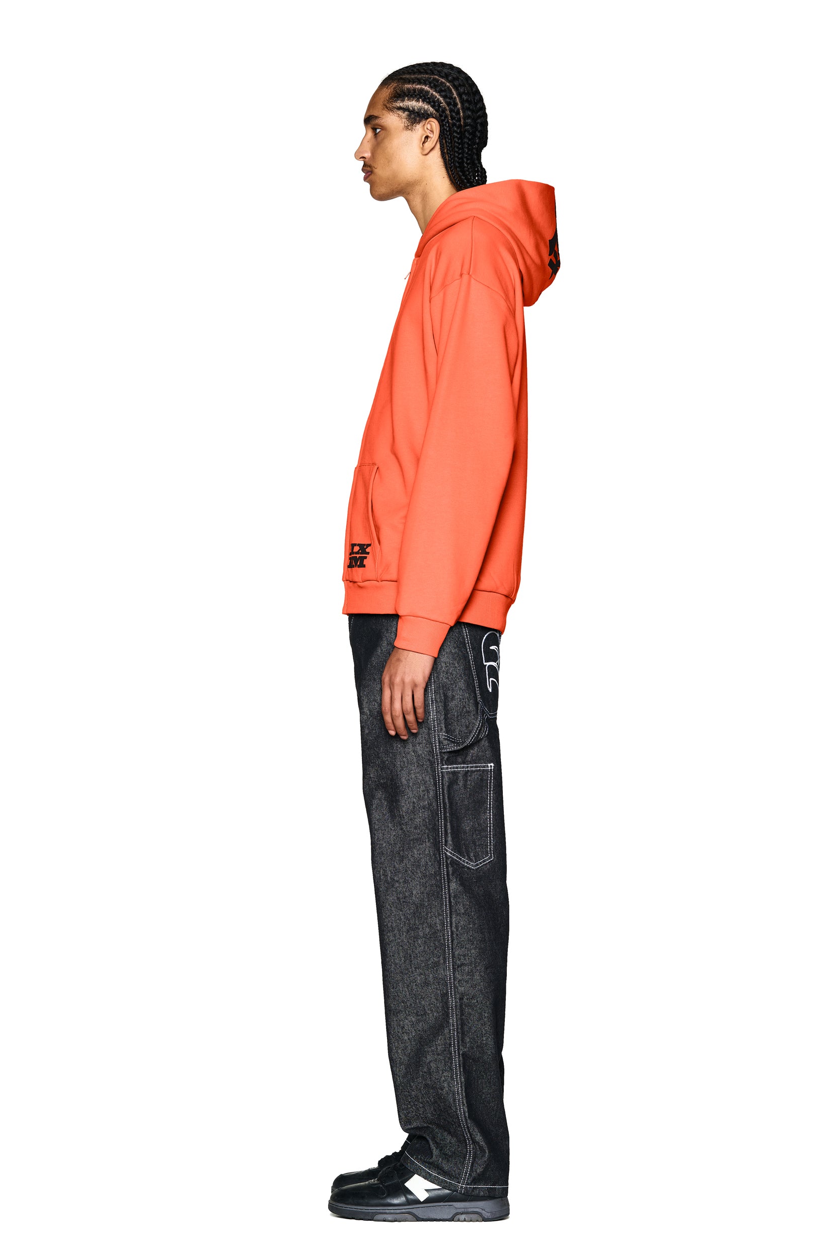 A person with braided hair stands in profile, wearing the 6PM LOGO ZIP HOODIE ORANGE, loose dark jeans with white stitching, and black sneakers against a plain white background.